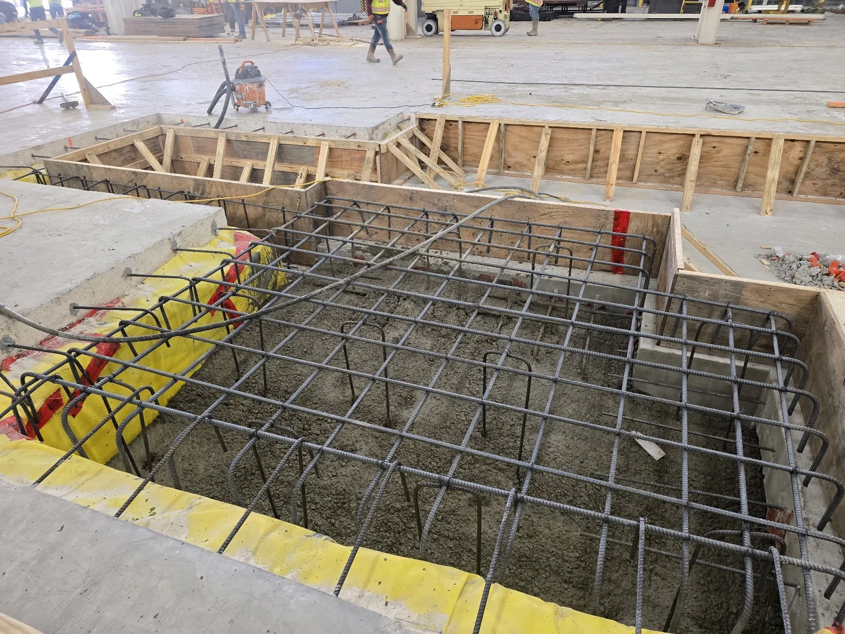 Construction site showing concrete foundation with rebar reinforcement and wooden formwork for a building.