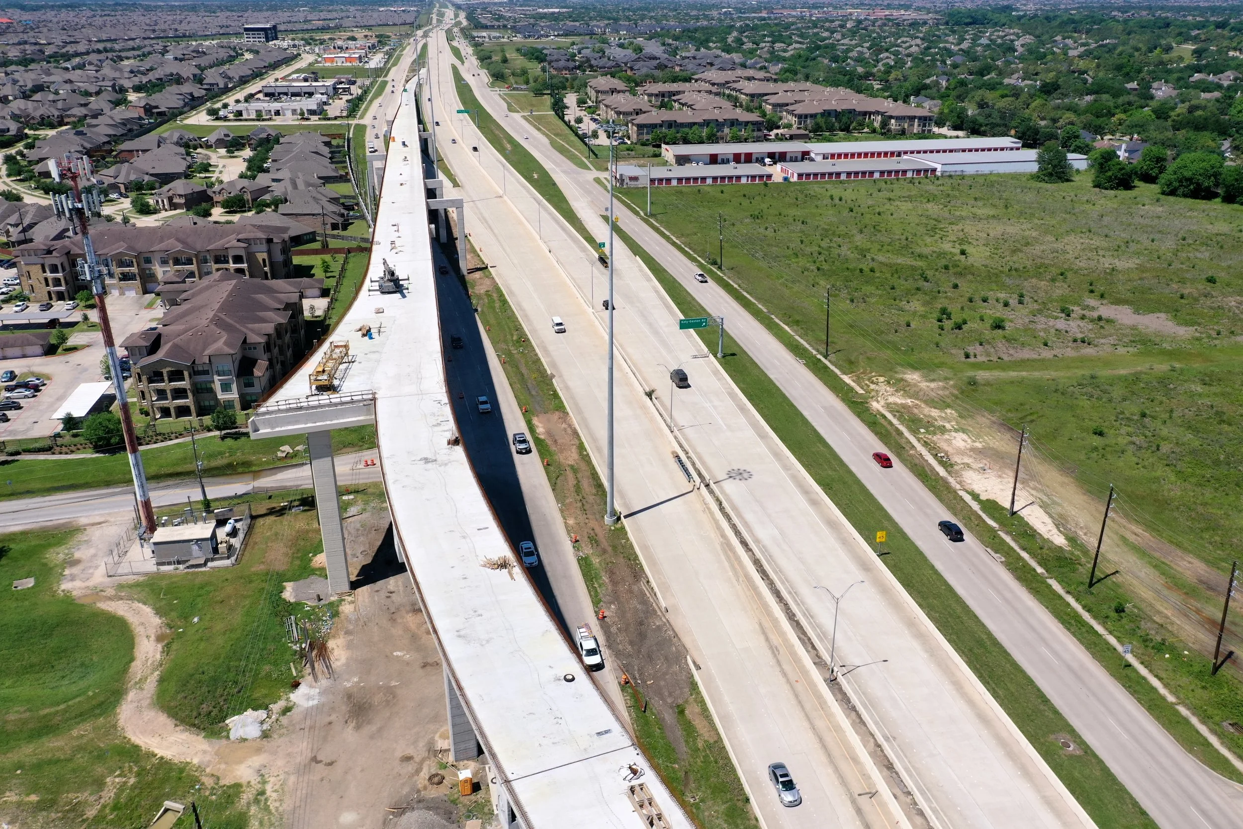 Aerial view of an ongoing construction of an elevated highway or bridge over a suburban area, with several vehicles on the partially built road and surrounding residential neighborhoods.