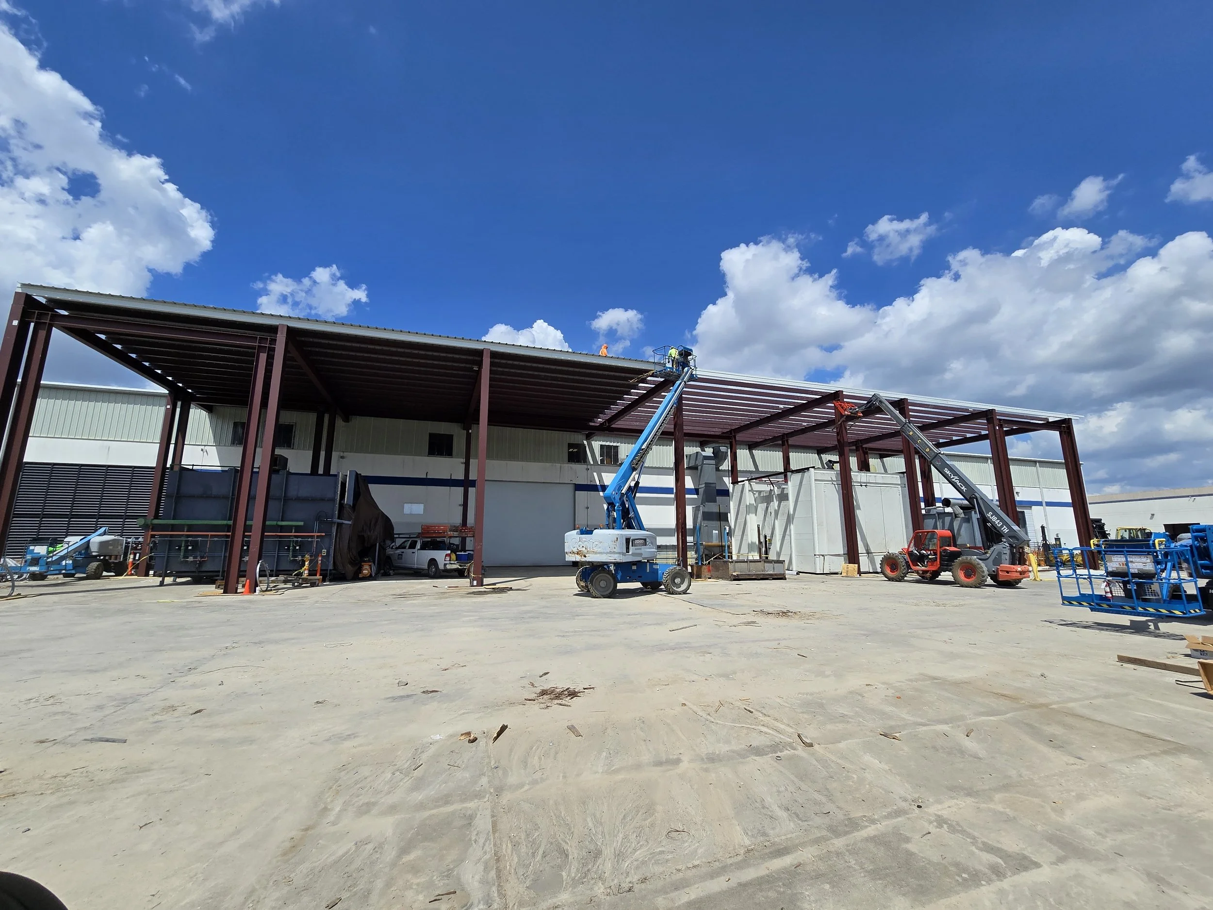 Construction site with workers on a roof using lifts, and heavy equipment including cranes, against a blue sky with clouds.