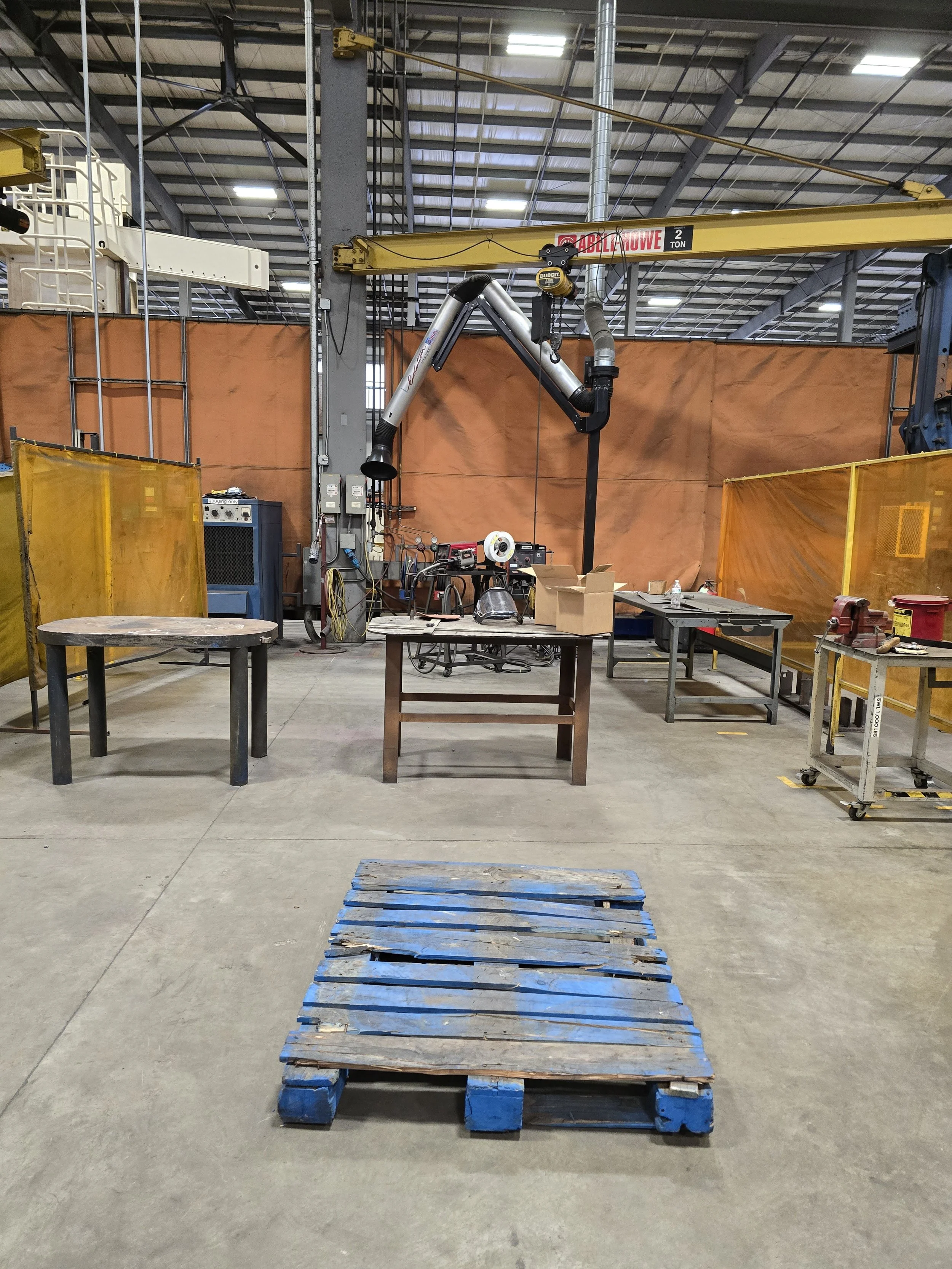 Industrial welding workshop with work tables, a robotic arm, and safety barriers, with a wooden pallet on the concrete floor.