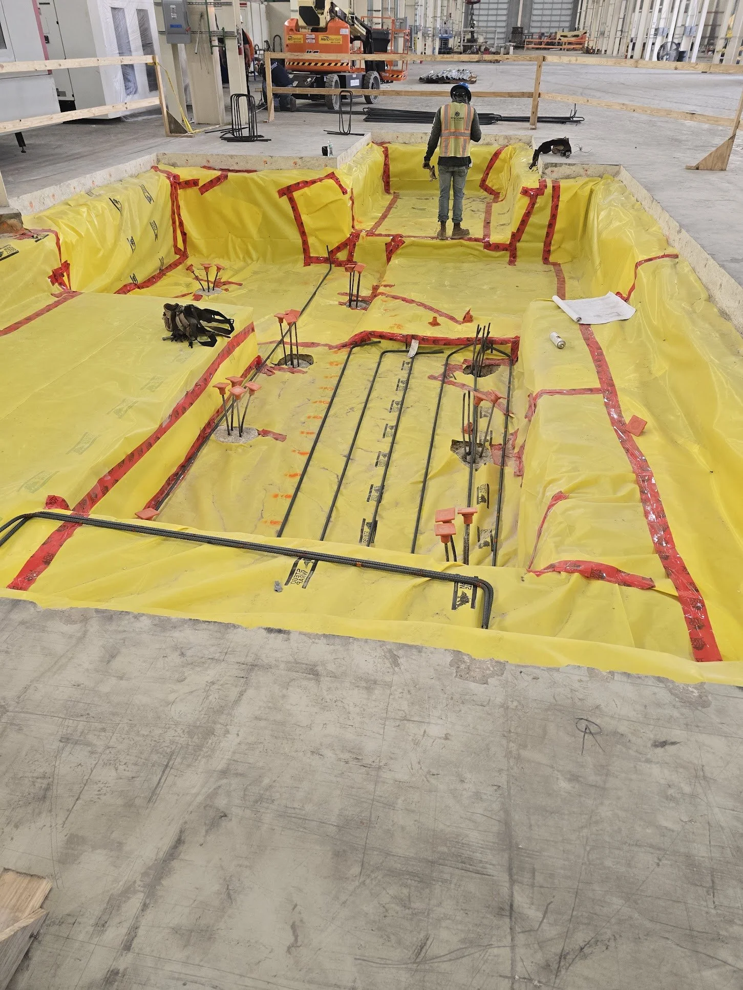 Construction site with a worker in a safety vest and helmet looking at a prepared floor area outlined with yellow plastic and red tape, with embedded pipes and rebar. Construction equipment and materials are visible in the background.