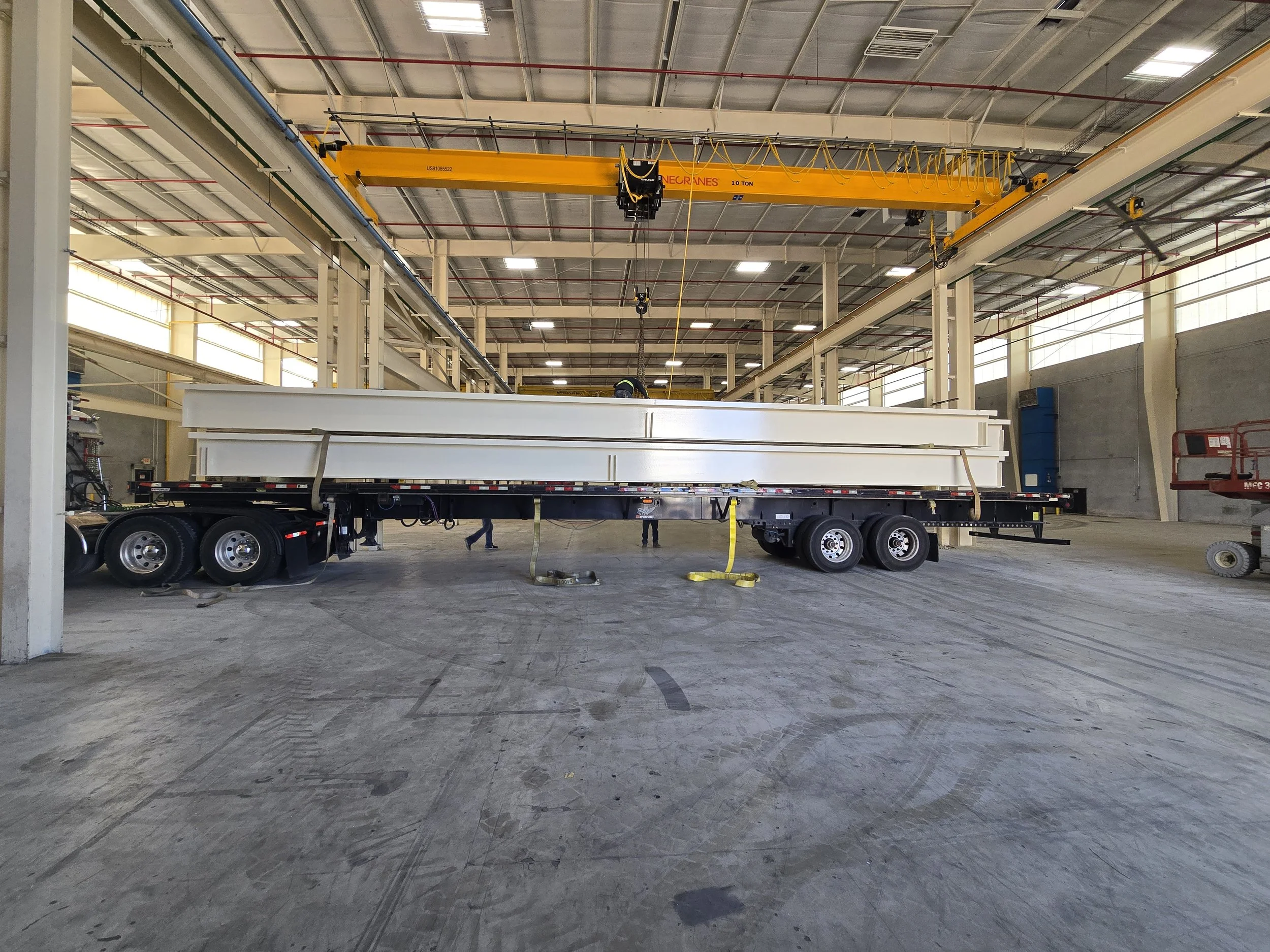 A large flatbed trailer carrying panels is inside an industrial warehouse with high ceilings, metal beams, and large windows, along with an overhead yellow crane.