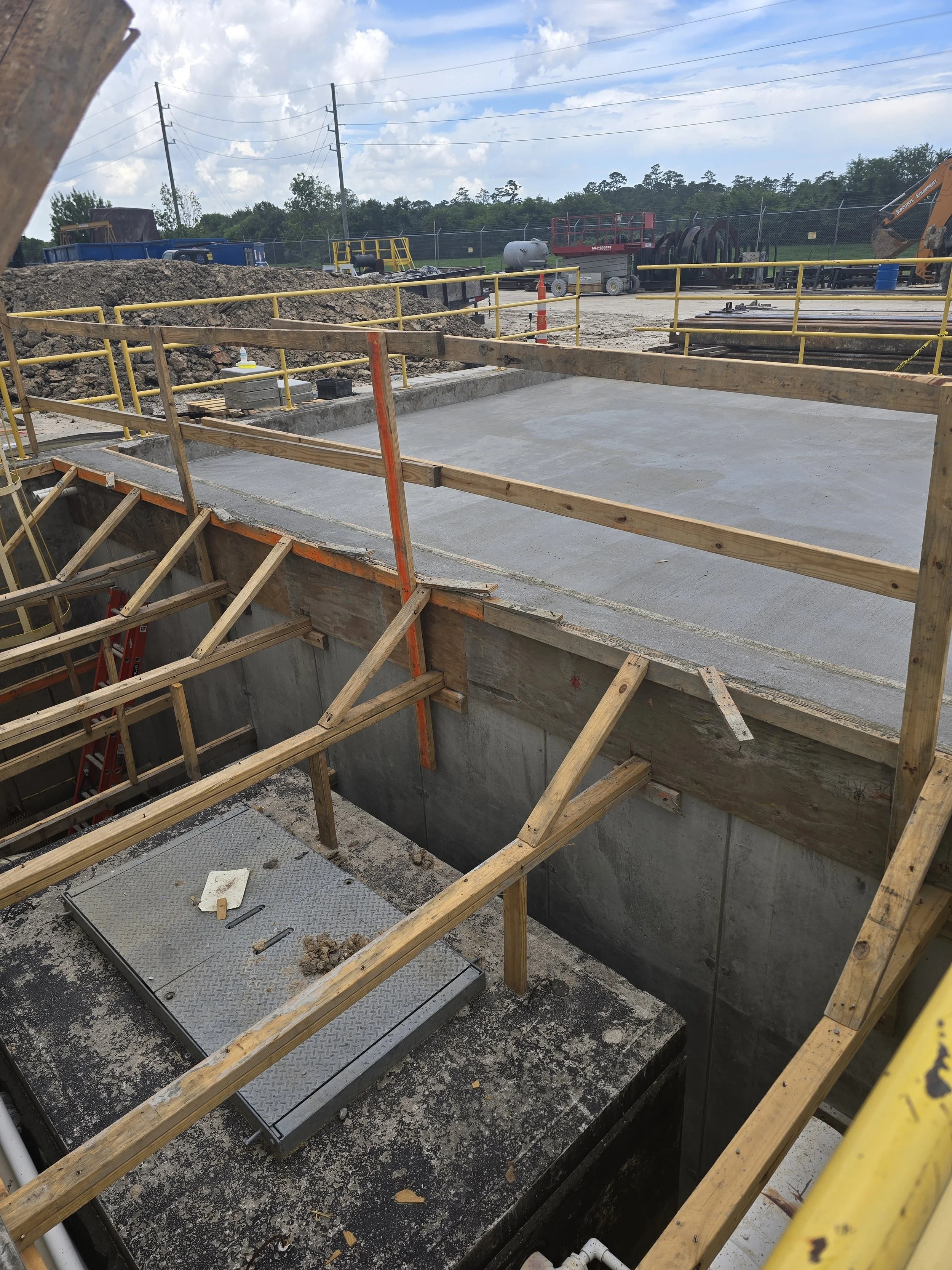 Construction site with concrete foundation, wooden scaffolding, safety railings, construction equipment, and a cloudy sky.