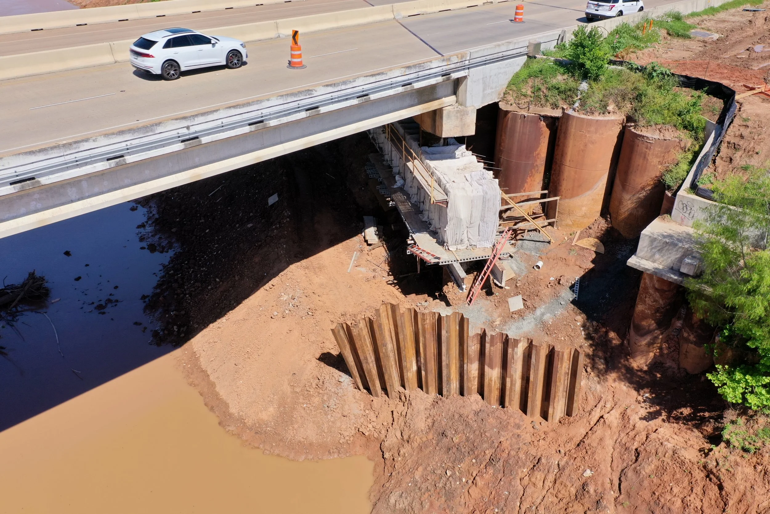 Under construction view of a bridge with construction materials, dirt, and support beams visible below, alongside vehicles on the road above.