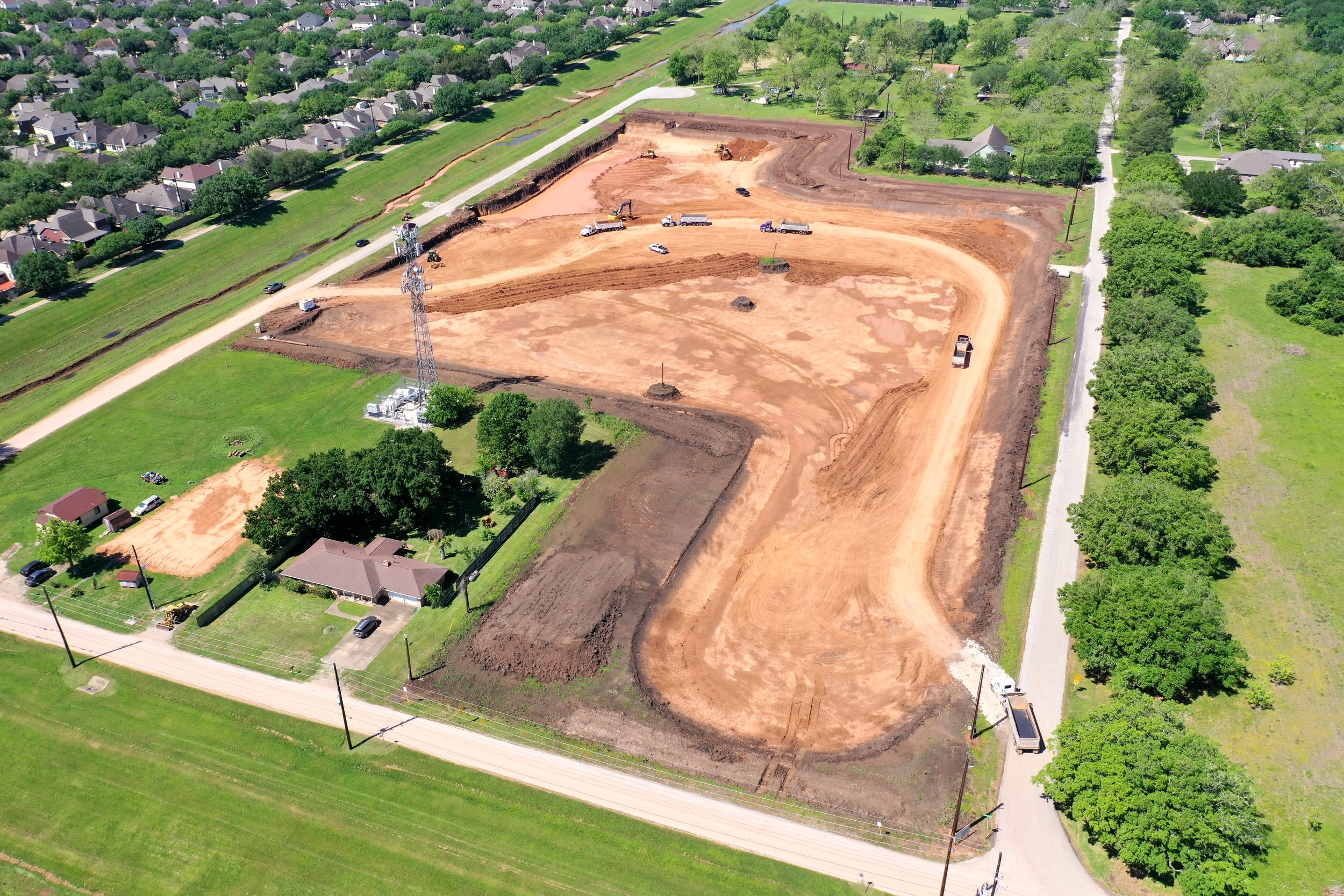 An aerial view of a construction site with cleared earth and several vehicles, bordered by residential houses and green trees.