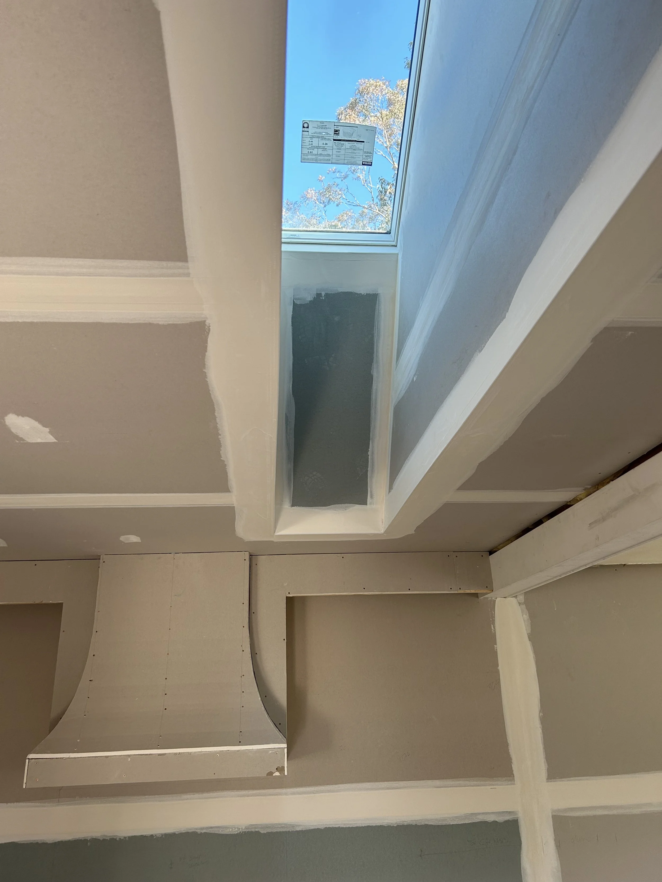 Interior of a house under construction, showing a rectangular window with a view of trees and blue sky, surrounded by drywall in progress.