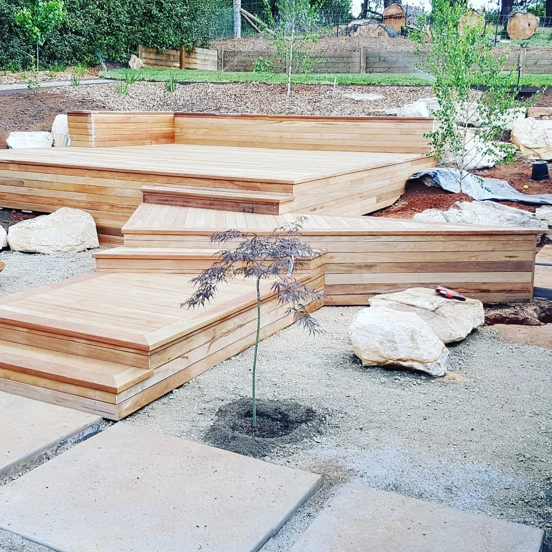 Wooden tiered garden steps or seating area with large rocks and small plants, in a landscaped yard.
