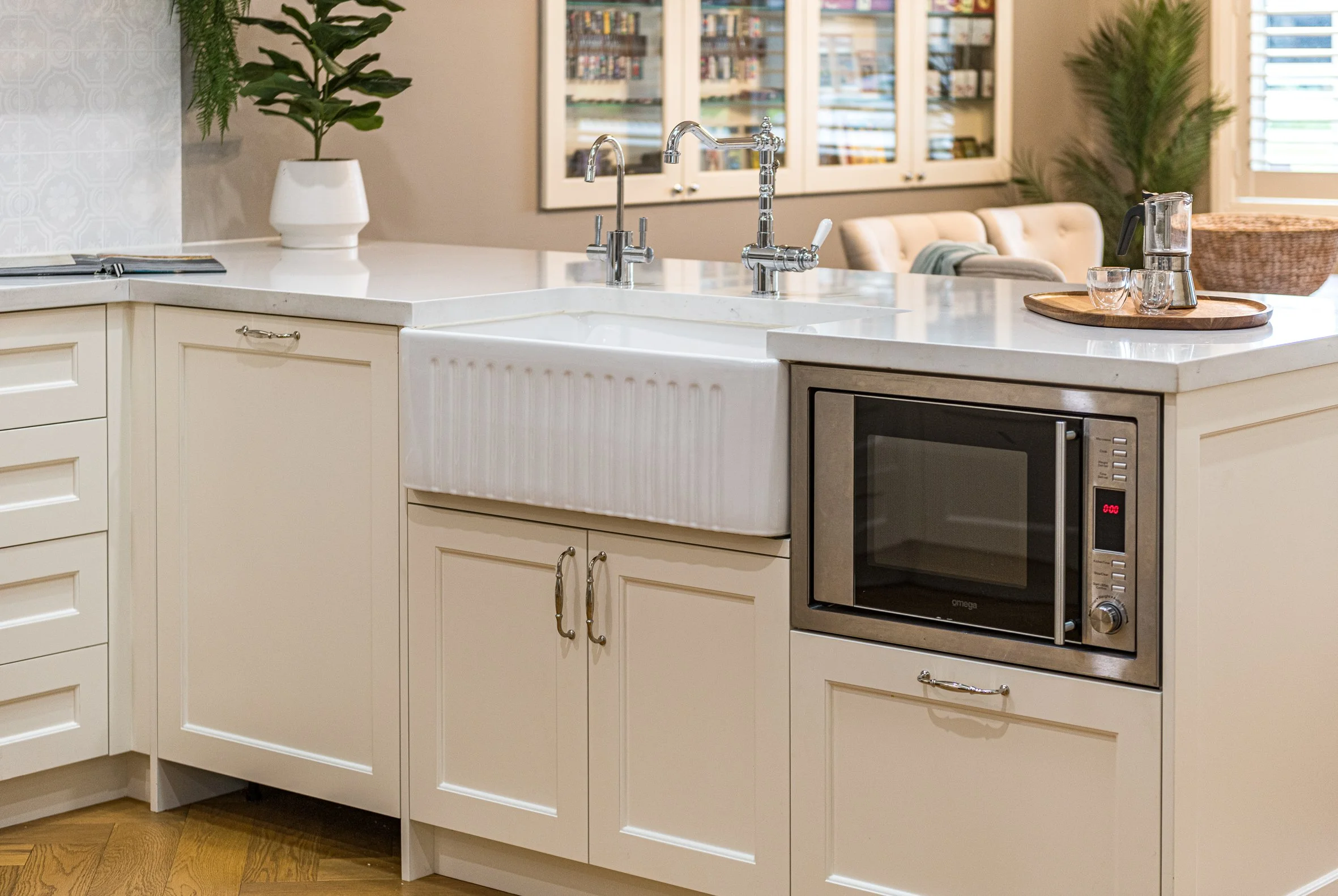A kitchen with a white farmhouse sink, a marble countertop, and a built-in microwave oven. There are two modern faucets over the sink, a potted plant, and a tray with a blender, glasses, and a wooden tray. In the background, there is a sitting area with a cream-colored sofa and creating a cozy space.