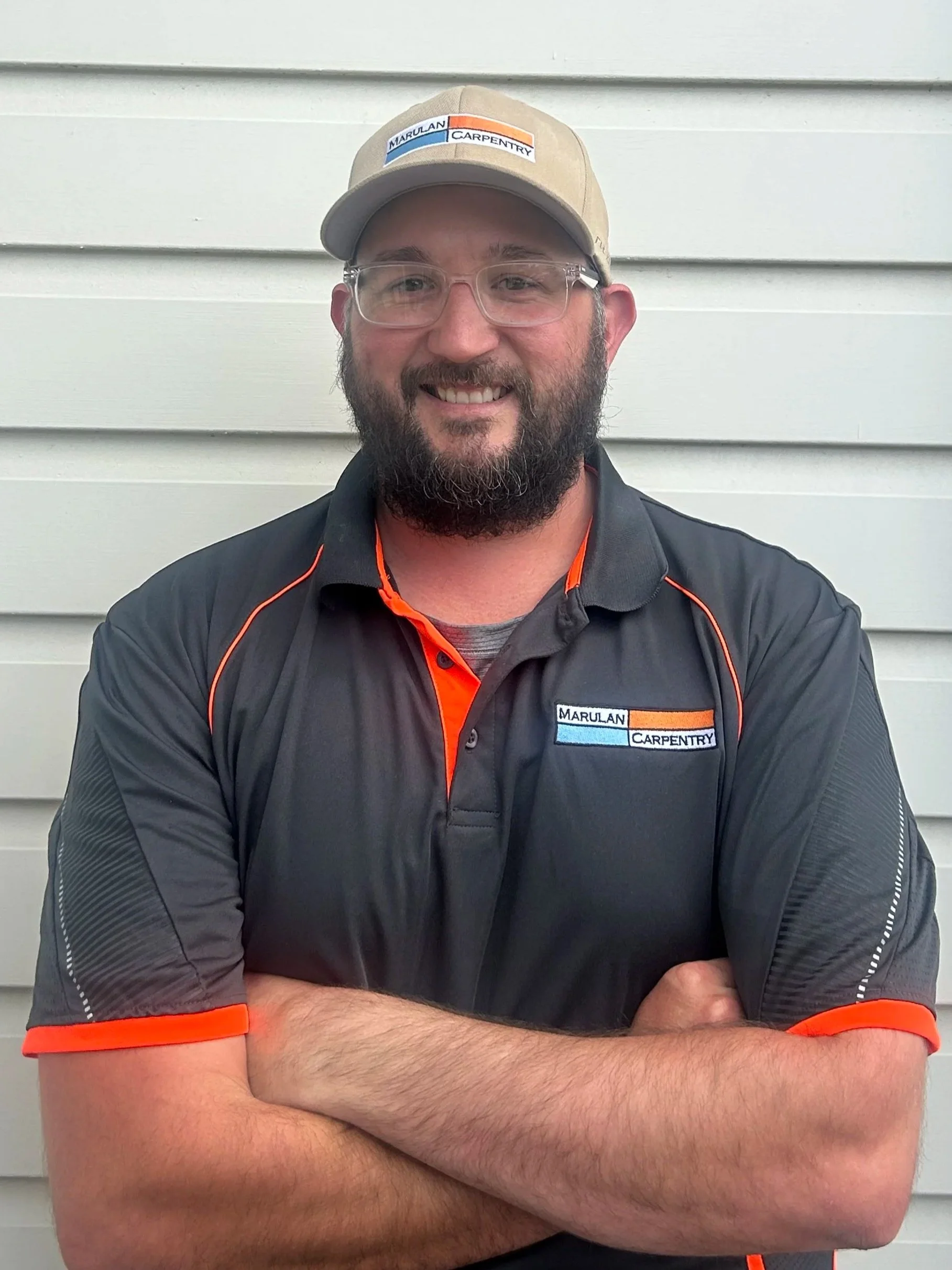 A smiling man with glasses, a beard, and crossed arms standing in front of a white siding wall, wearing a baseball cap and a black Marulan Carpentry shirt.