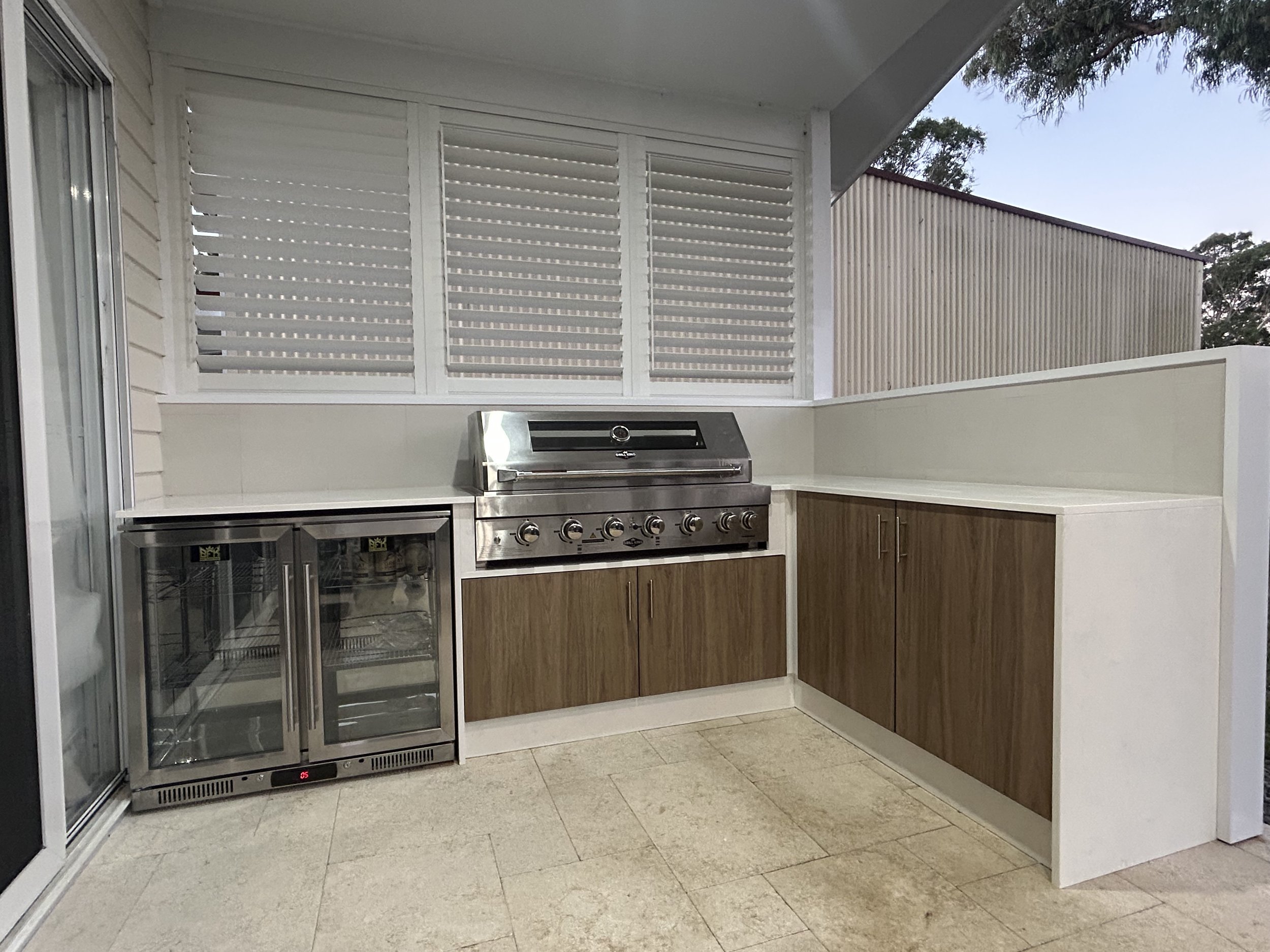 Outdoor kitchen with stainless steel grill, mini refrigerator, wooden cabinets, white countertop, and privacy shutters on the wall.