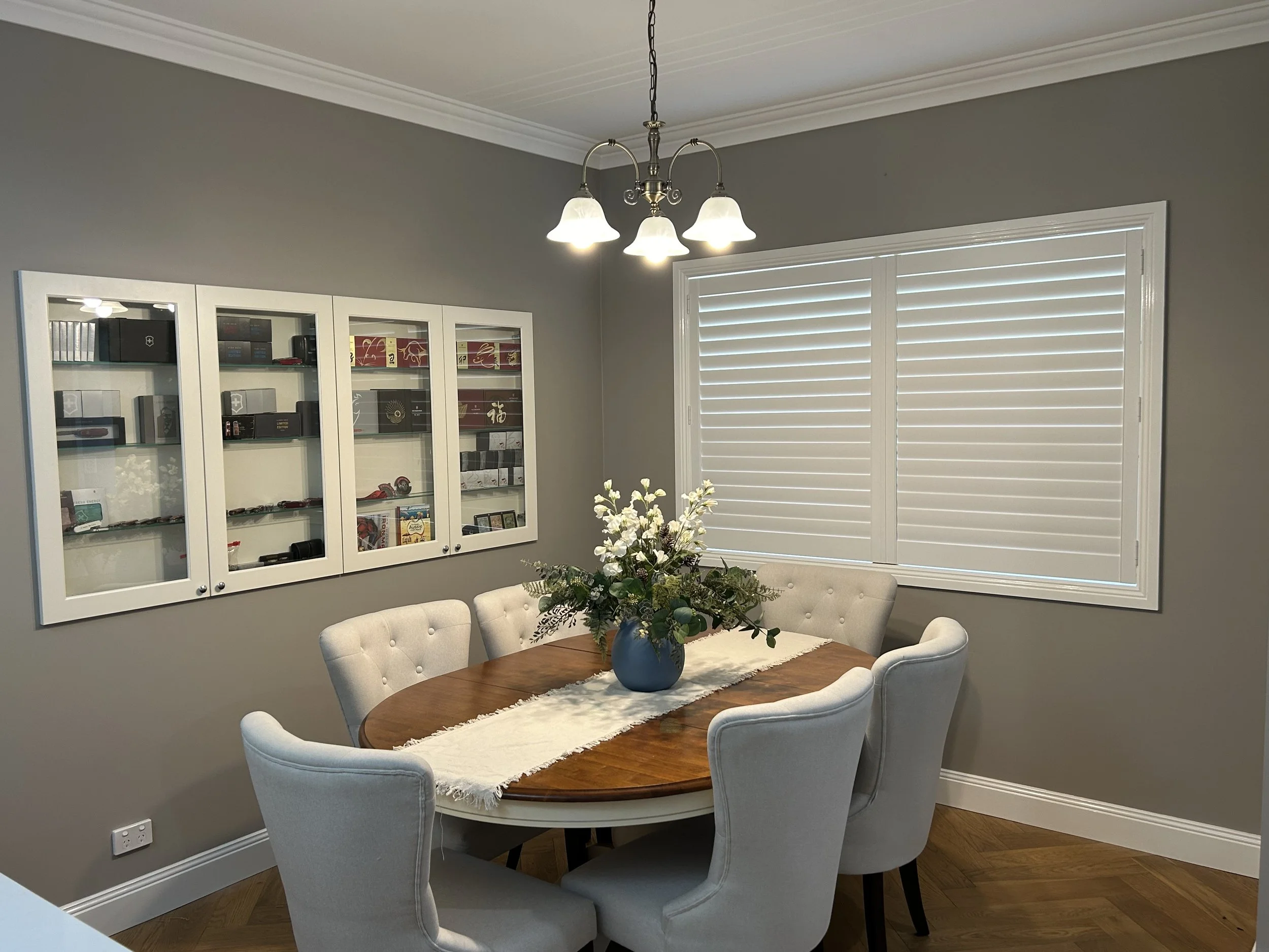 Modern dining room with a wooden table, six upholstered chairs, a vase of flowers, a chandelier, and a wall display case with collectibles, next to windows with closed white blinds.
