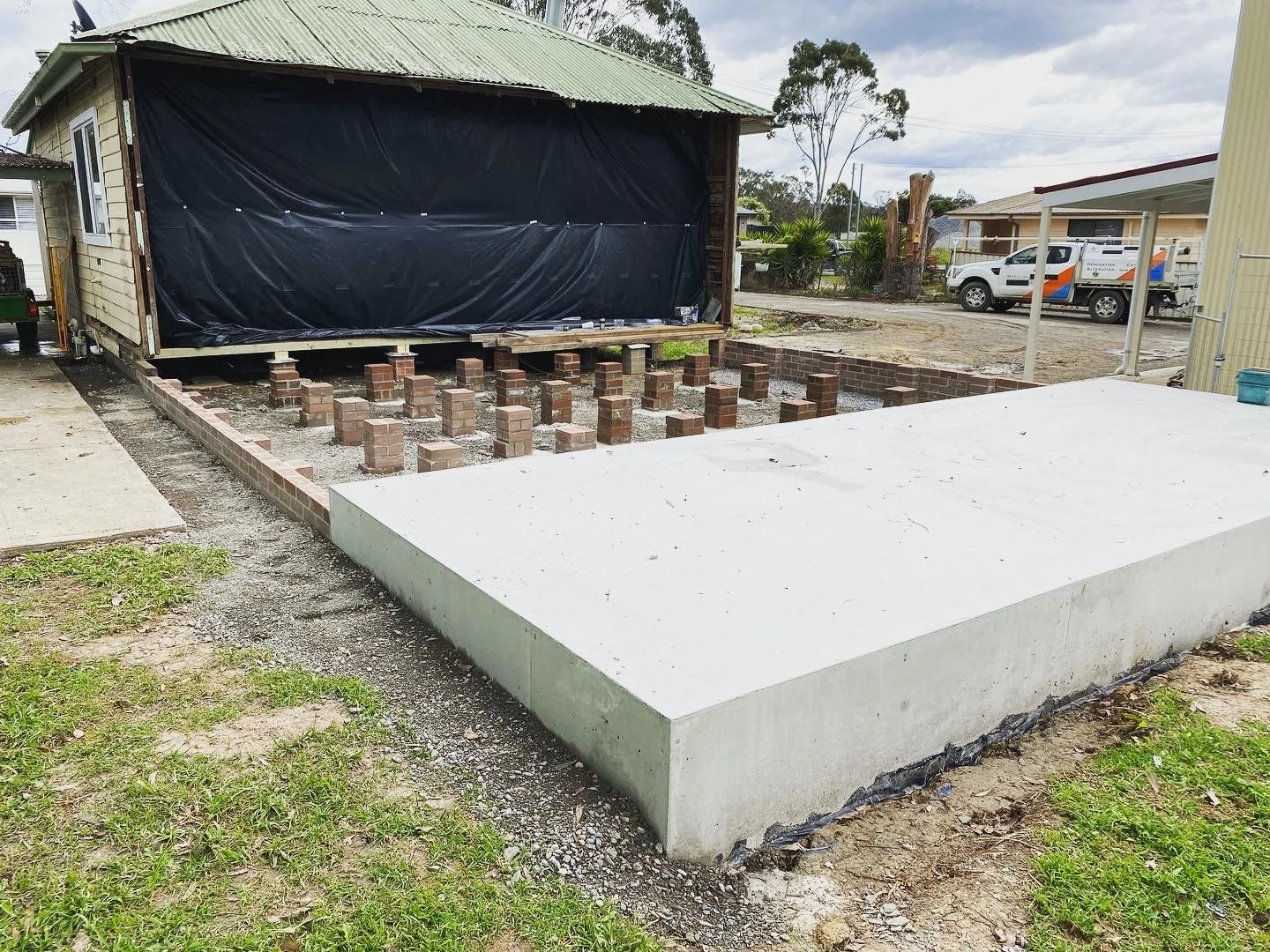 Construction site with brick foundation and concrete slab, house in progress, parked vehicles in background.