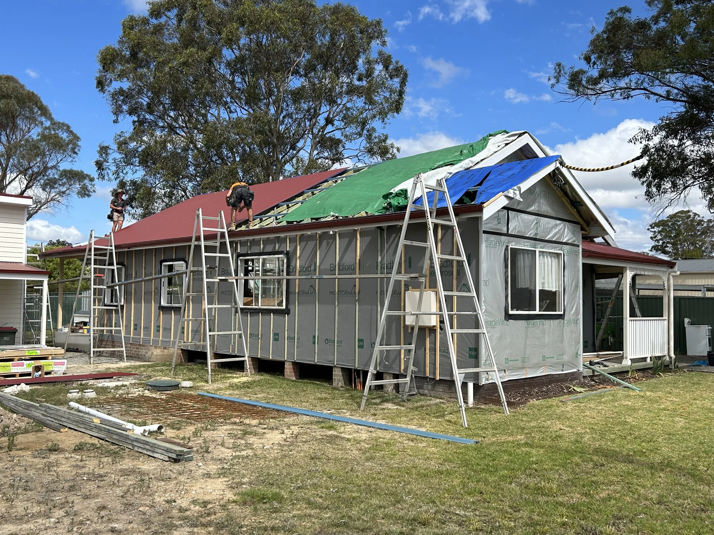 House under construction with workers installing a new red roof, some siding still being added, and ladders leaning against the house, surrounded by trees and blue sky.