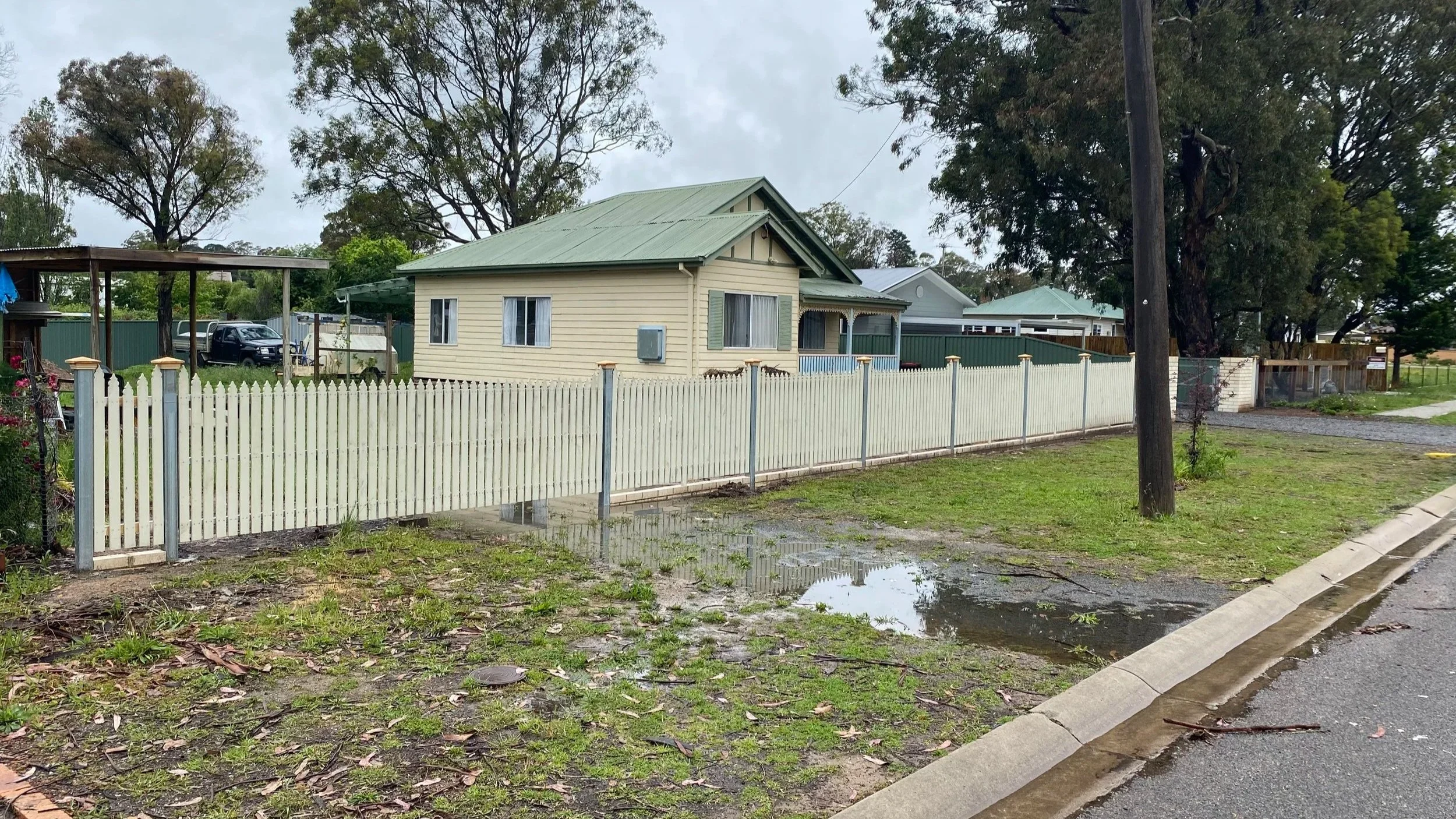 A backyard with a cream-colored house, white picket fence, potted plants, a carport, and trees in a neighborhood.