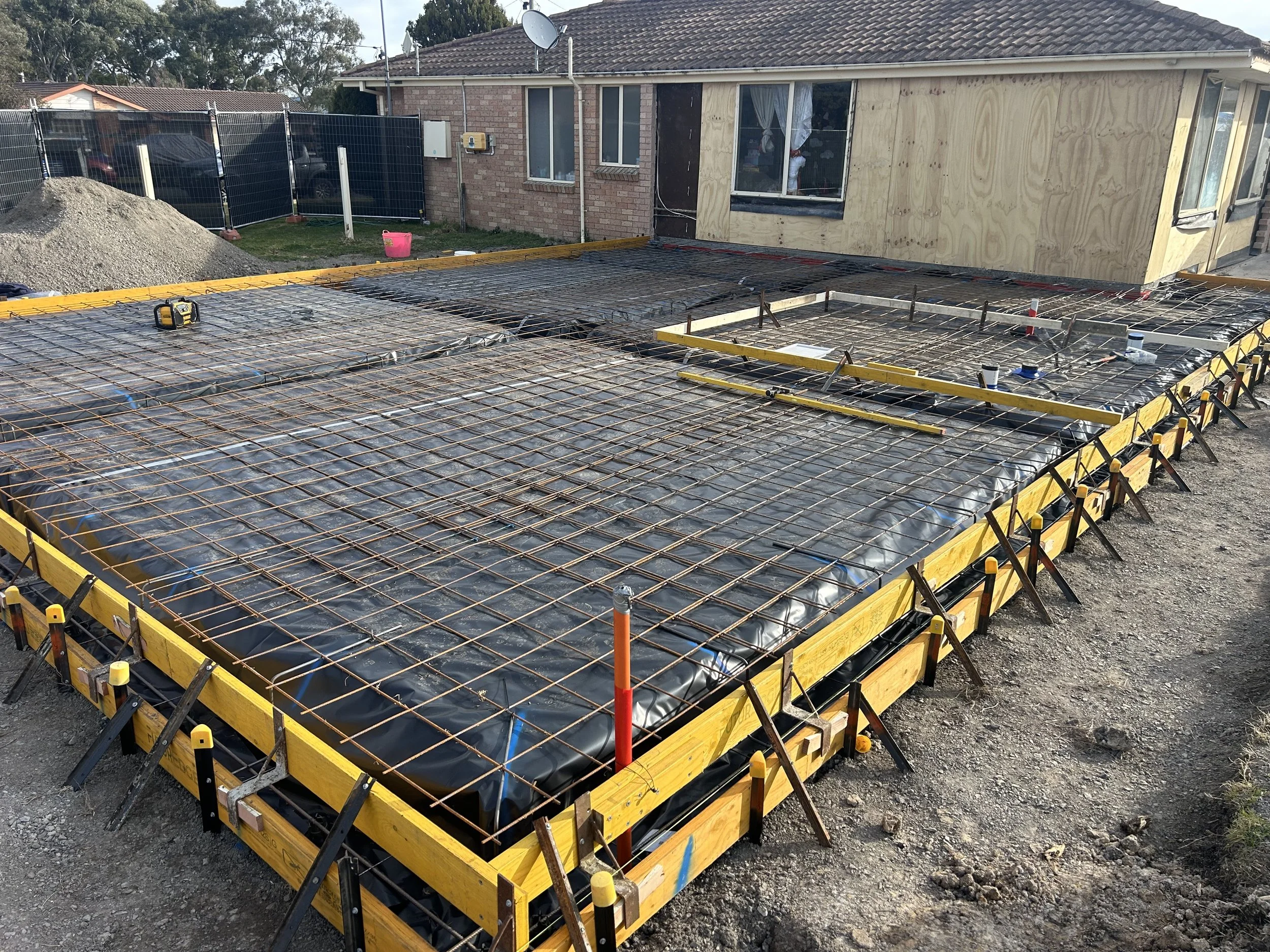 Construction site with rebar grid and black plastic sheeting, preparing for concrete pouring outside a brick house with unfinished walls and a small extension.