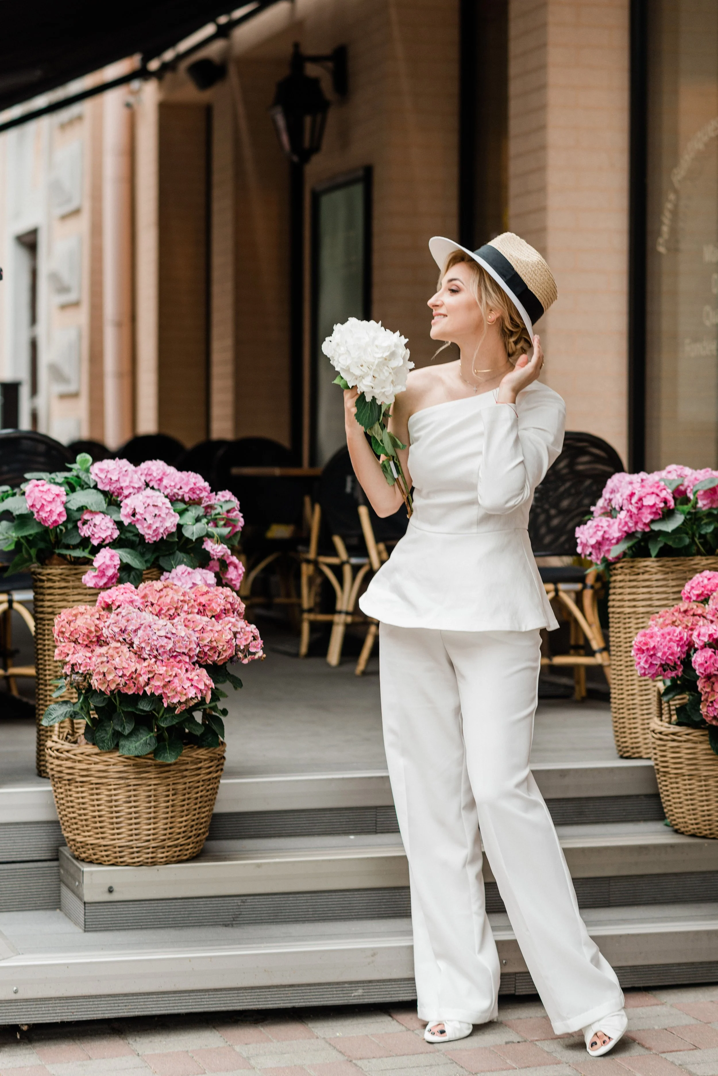 A woman dressed in white with a wide-brimmed hat, holding a bouquet of white flowers, standing in front of pink hydrangeas in woven baskets outside a building.