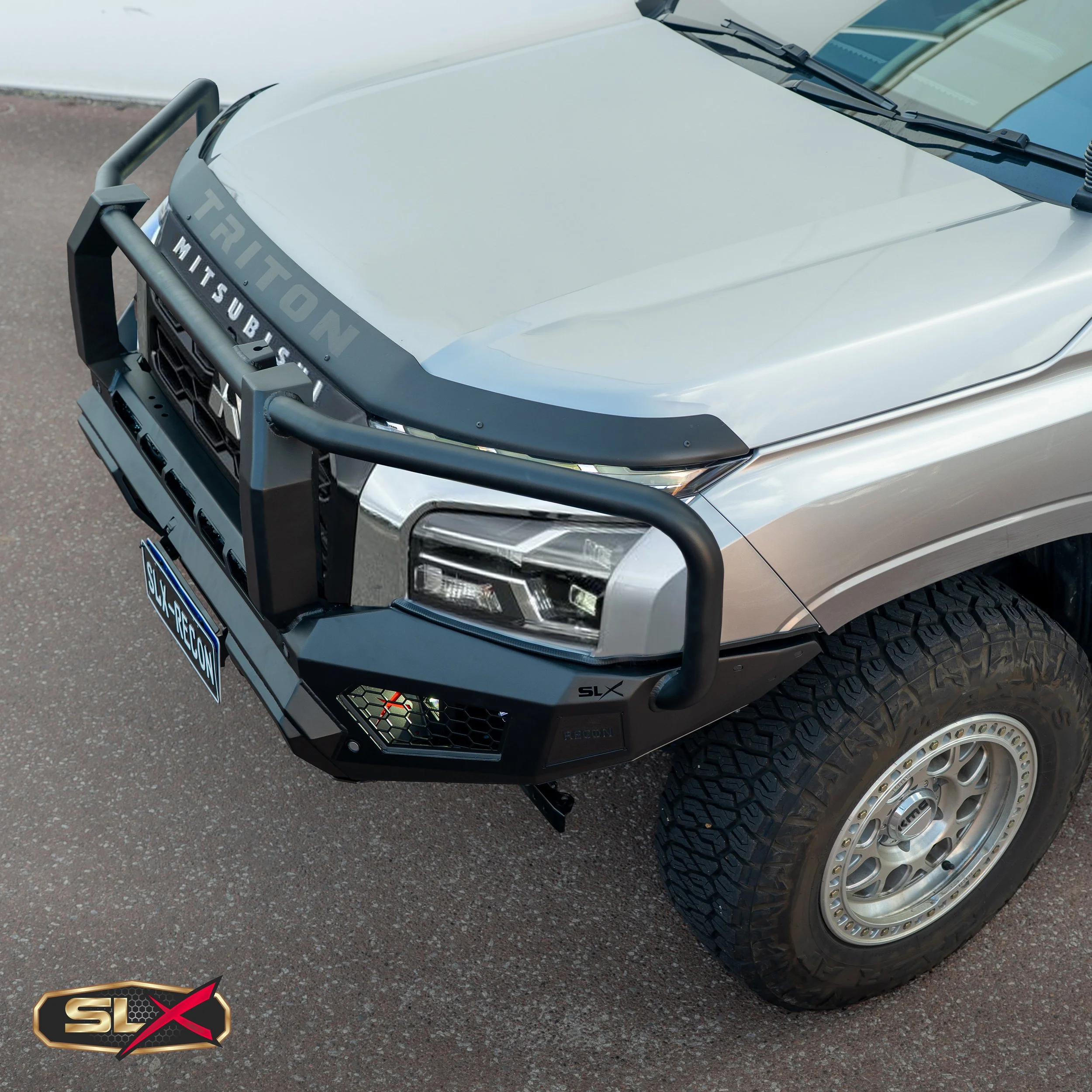 Close-up of the front of a silver Mitsubishi truck with a black reinforced grill guard bearing the word 'TRITON'. The truck has large off-road tires, a black bumper, and a black license plate with the word 'Slyfoxcon'.