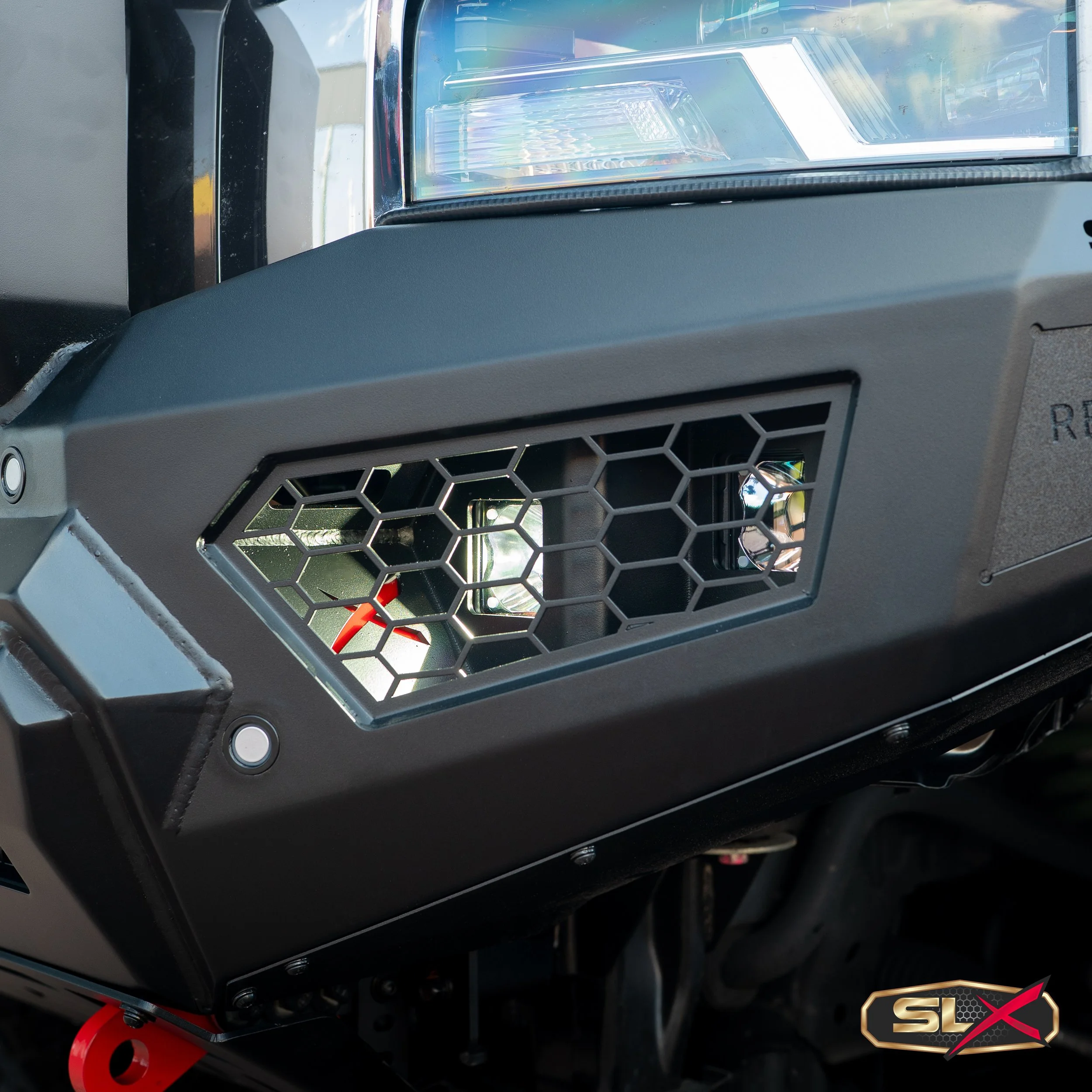 Close-up image of a vehicle's front end, showing a modern headlight and a black grill with honeycomb pattern, with the SLX logo in the lower right corner.