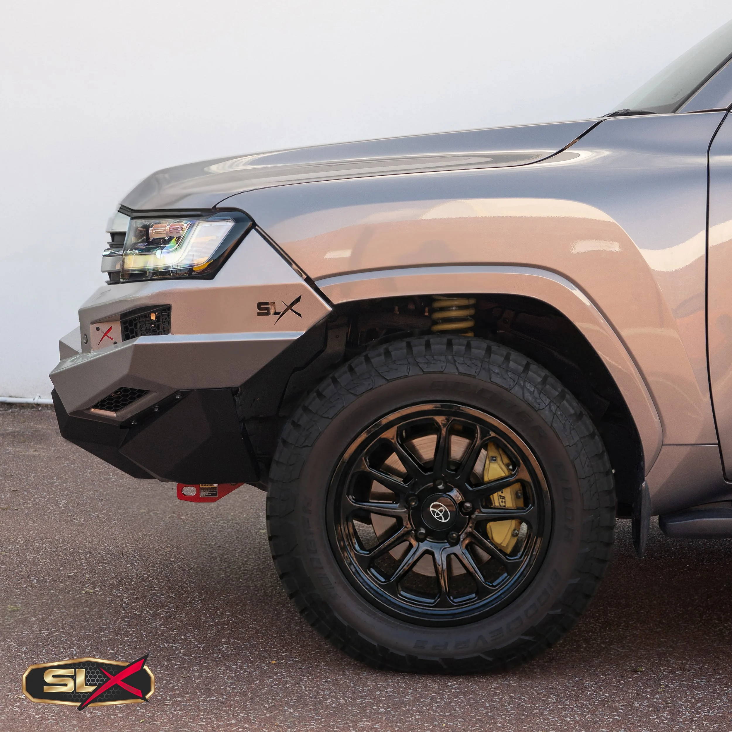 Close-up of a gray Toyota Tacoma pickup truck's front left side with off-road tires, black wheels, and a black bumper. The truck has a decal with 