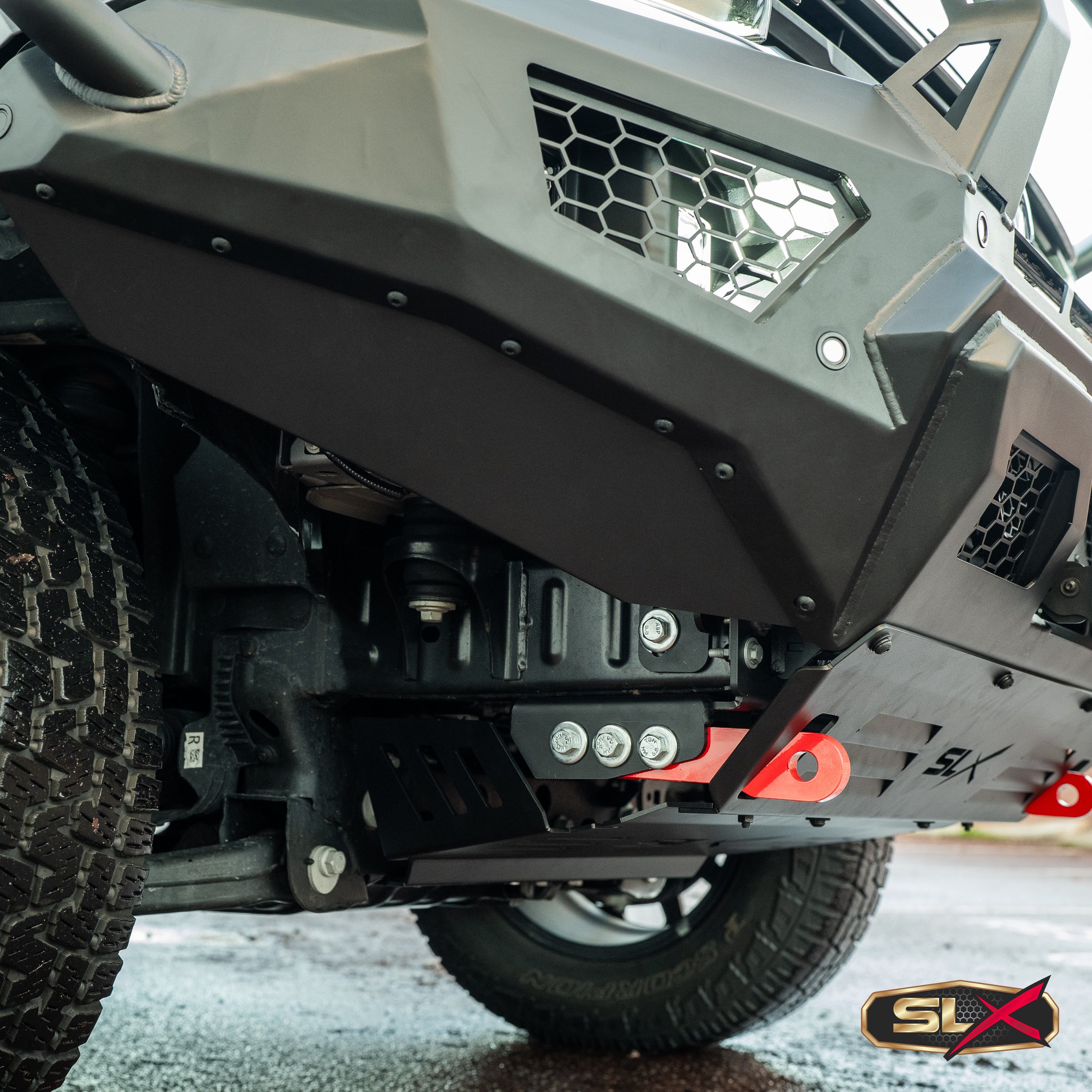 Close-up of armored off-road vehicle's front undercarriage with custom skid plate, rugged tire, and logo in bottom right corner.