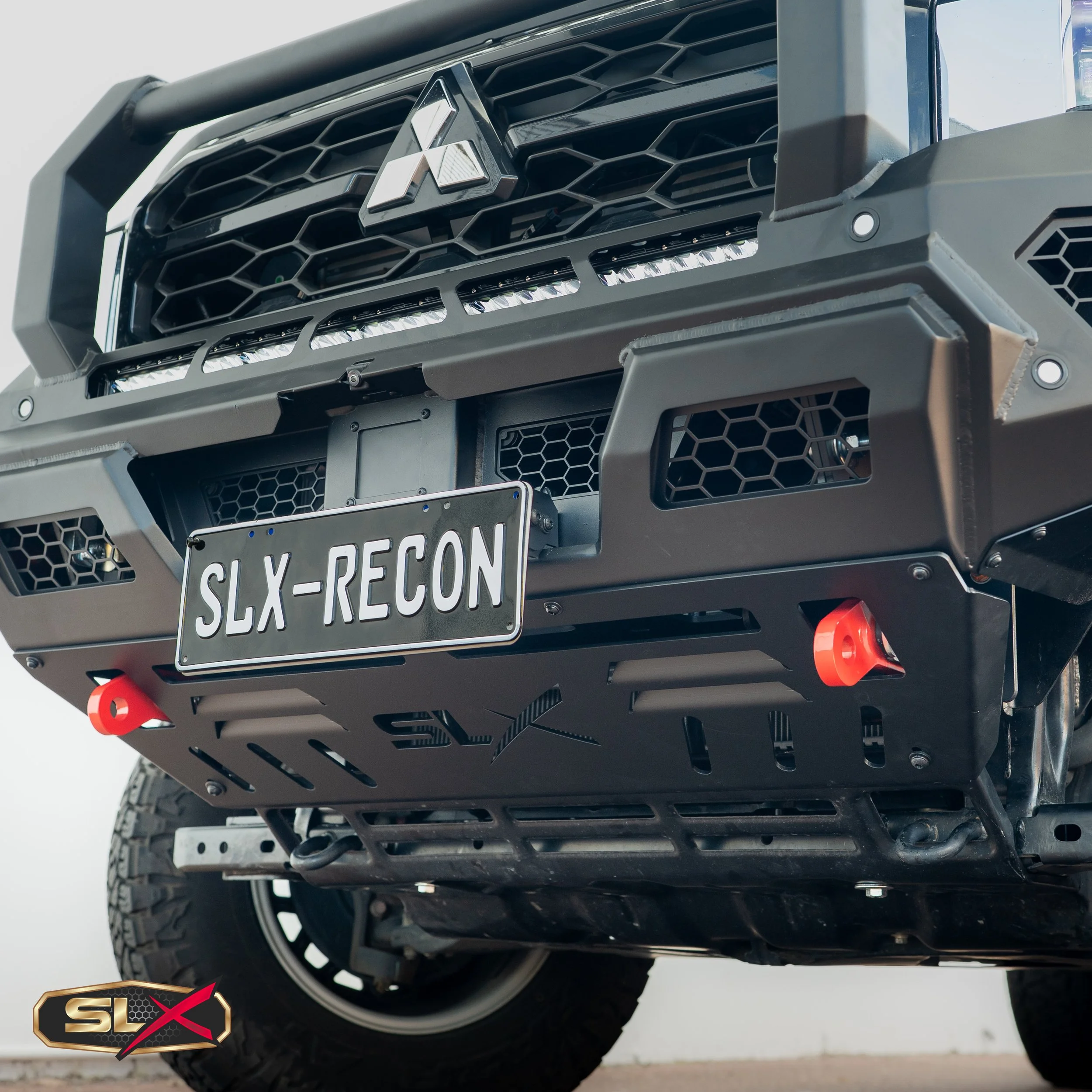 Close-up of the front bumper and grille of a black off-road vehicle with a license plate reading 'SLX-RECON'. The vehicle has a Mitsubishi emblem on the grille, off-road lights, and a skid plate with the 'SLX' logo.