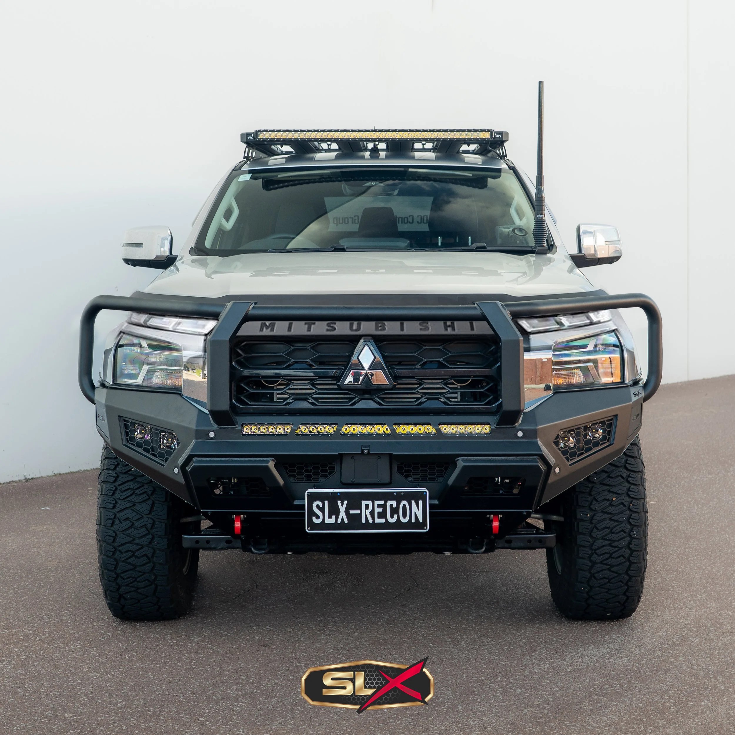 Front view of a modified Mitsubishi off-road vehicle with a custom bumper, grille, and a license plate reading 'SLX-RECON' parked on asphalt against a white wall.