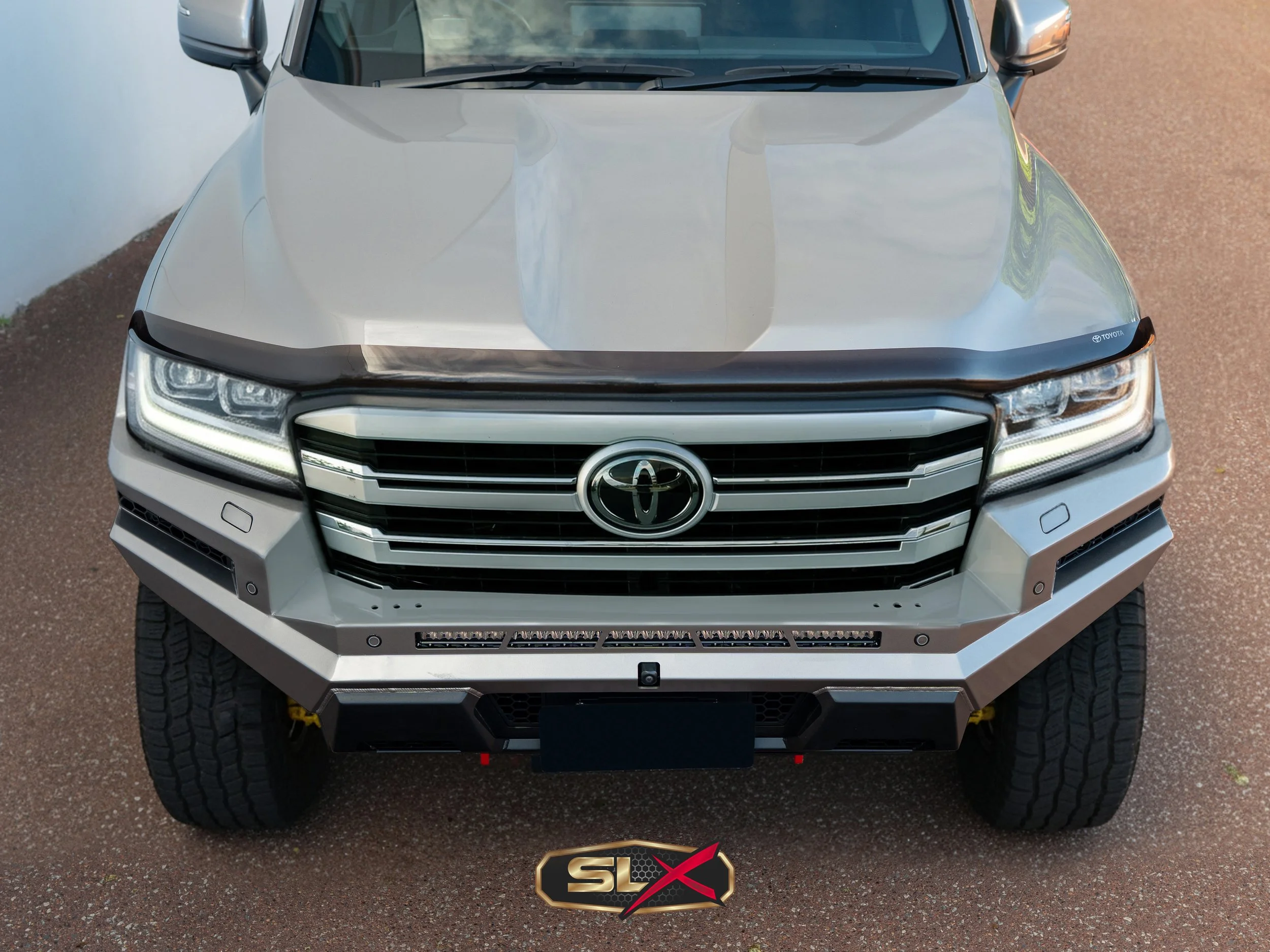 Front view of a silver Toyota SUV with a custom aluminum front bumper, LED light bar, and black hood protection, parked on a reddish-brown paved surface.
