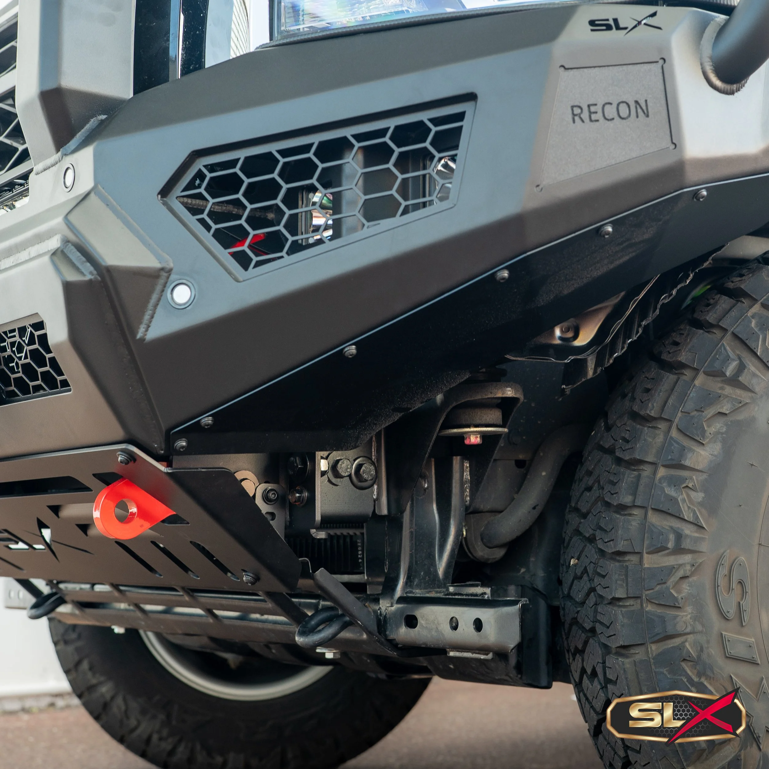 Close-up of the front underside of a rugged off-road vehicle, showing a black skid plate, large tire, and a grille with hexagonal openings. The vehicle has a label with the text 'RECON' and a badge with the logo 'SLX'.