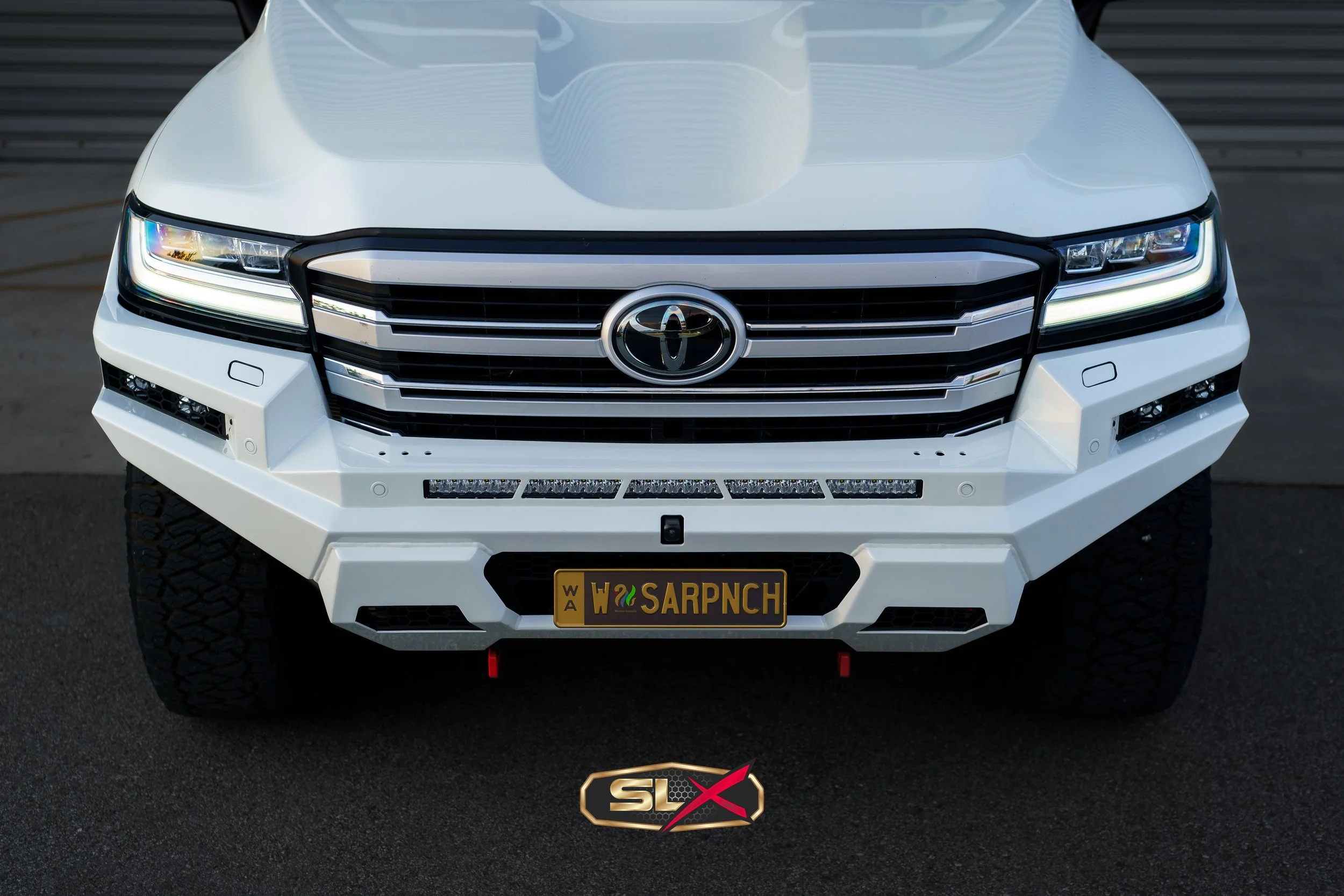 Front view of a white modified Toyota truck with black accents, custom license plate reading 'W SARP NCH', and a logo saying 'SLX' beneath it, parked on a concrete surface.
