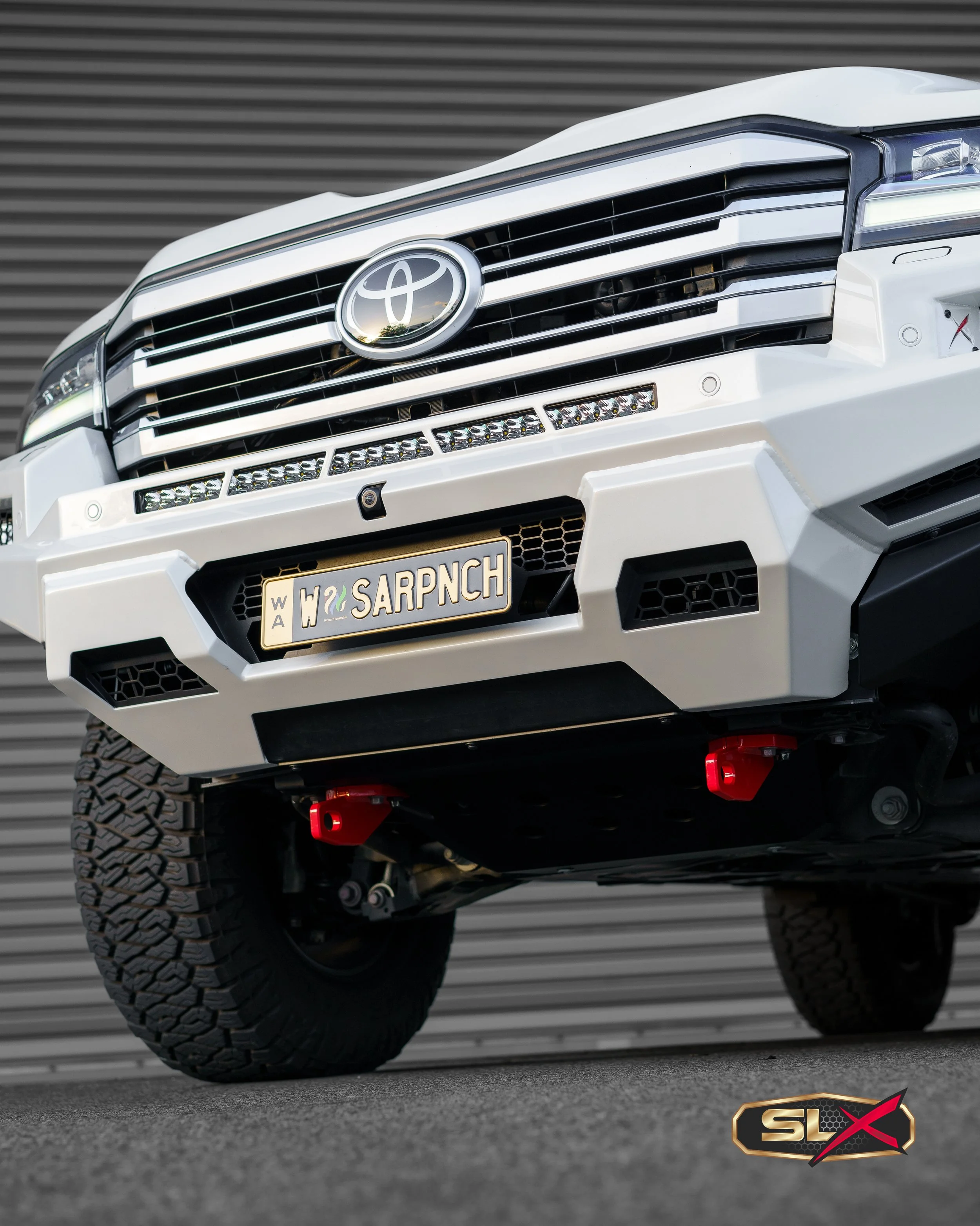 Close-up of a white Toyota SUV front with off-road tires, custom front bumper, red tow hooks, and a custom license plate that reads 'SARPUNCH', against a gray metal background.
