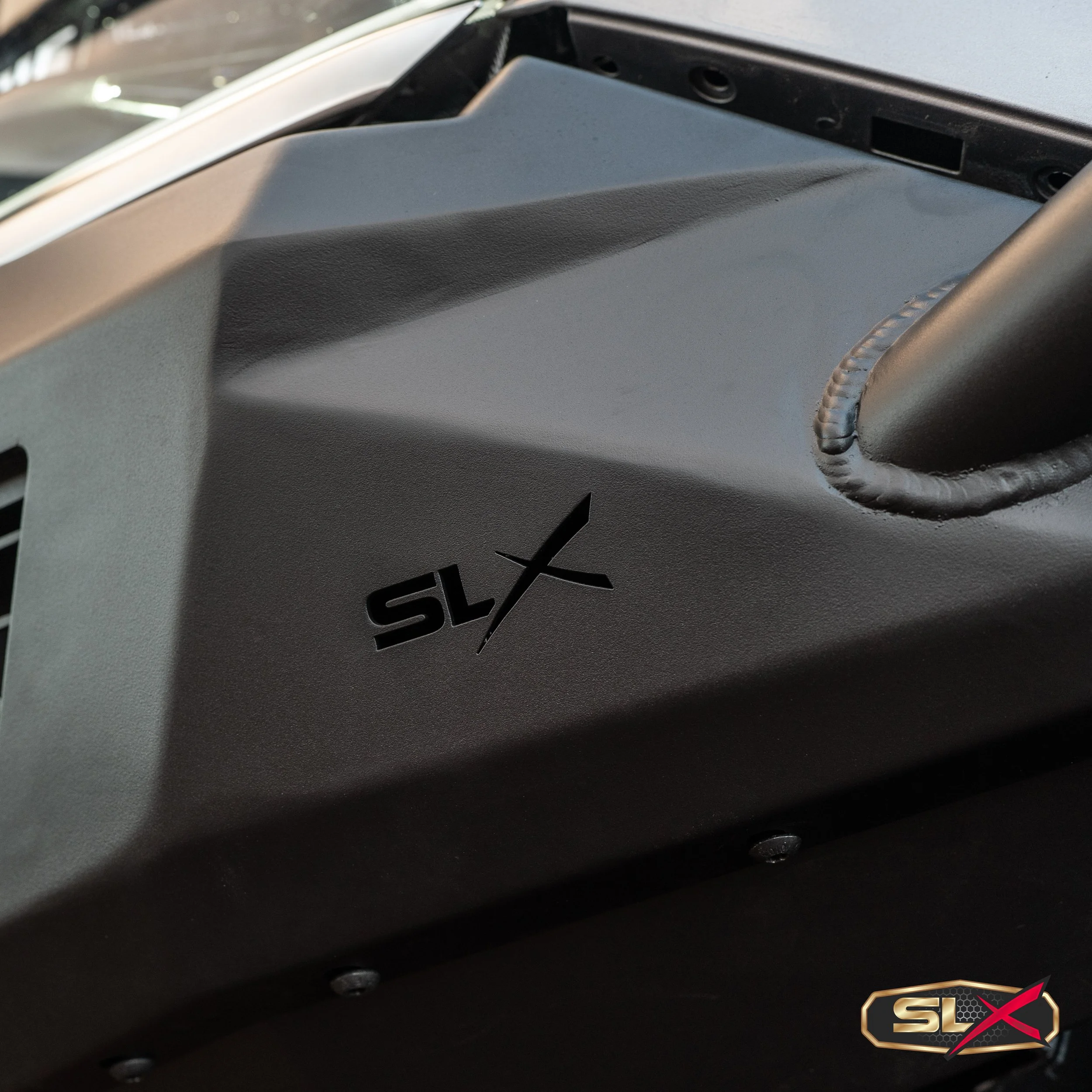 Close-up of a textured black surface on a vehicle with a cutout logo reading 'SLX' and a small, separate logo in the bottom right corner.