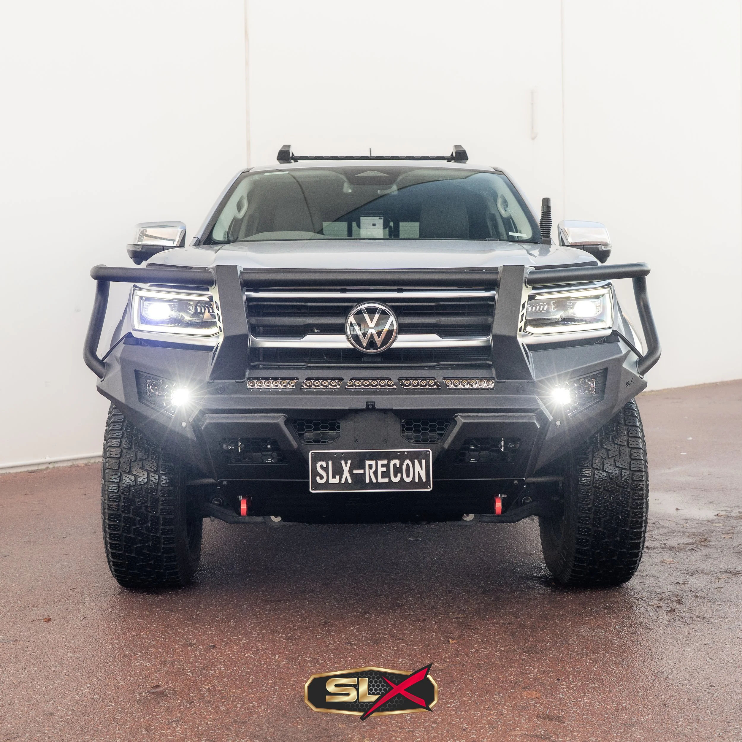 Front view of a black Volkswagen off-road vehicle with a large bumper guard, LED headlights, and a license plate that reads 'SLX-RECON' in a warehouse or garage setting.