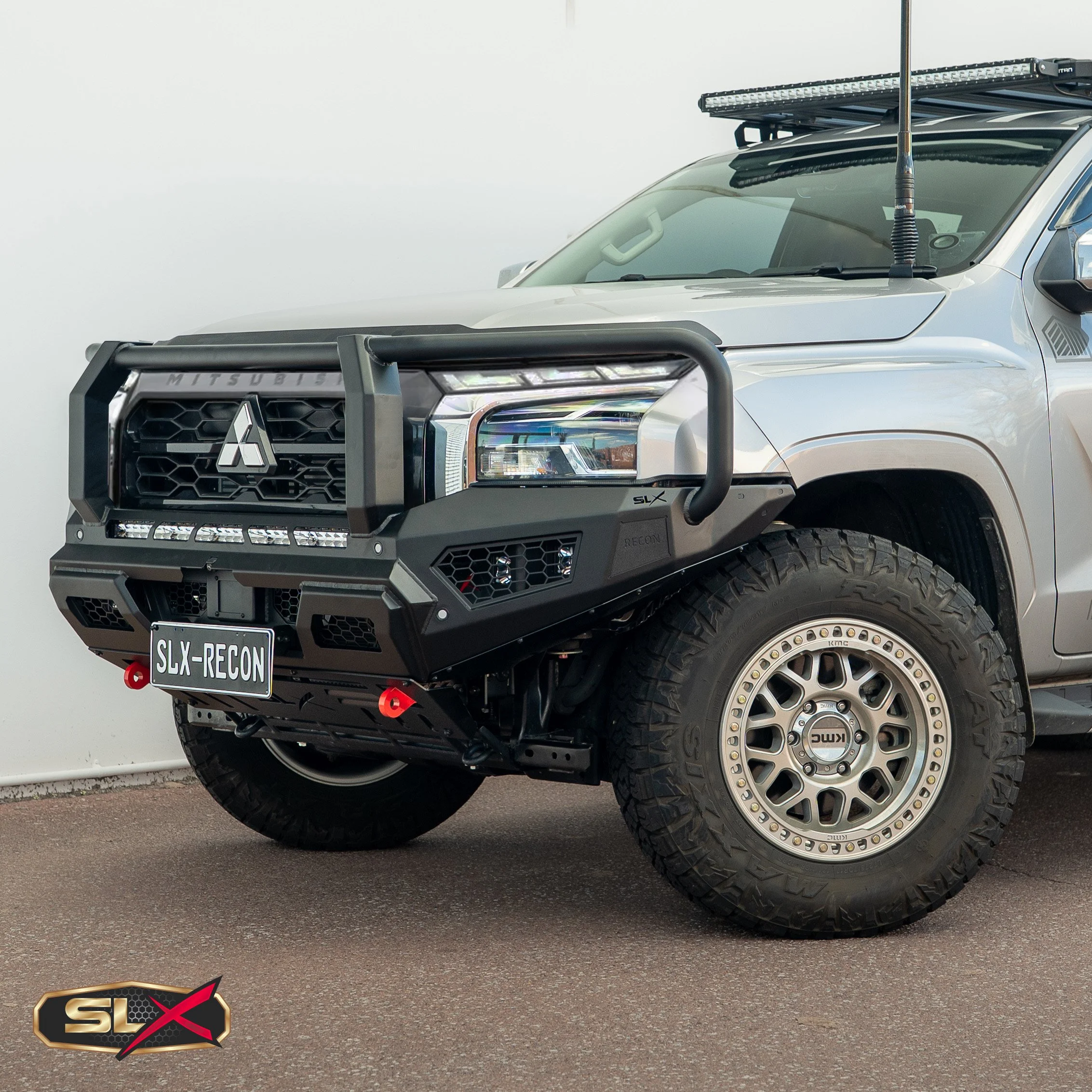 Silver off-road vehicle with a black bumper guard, large tires, and a light bar on top, parked indoors against a plain white wall.
