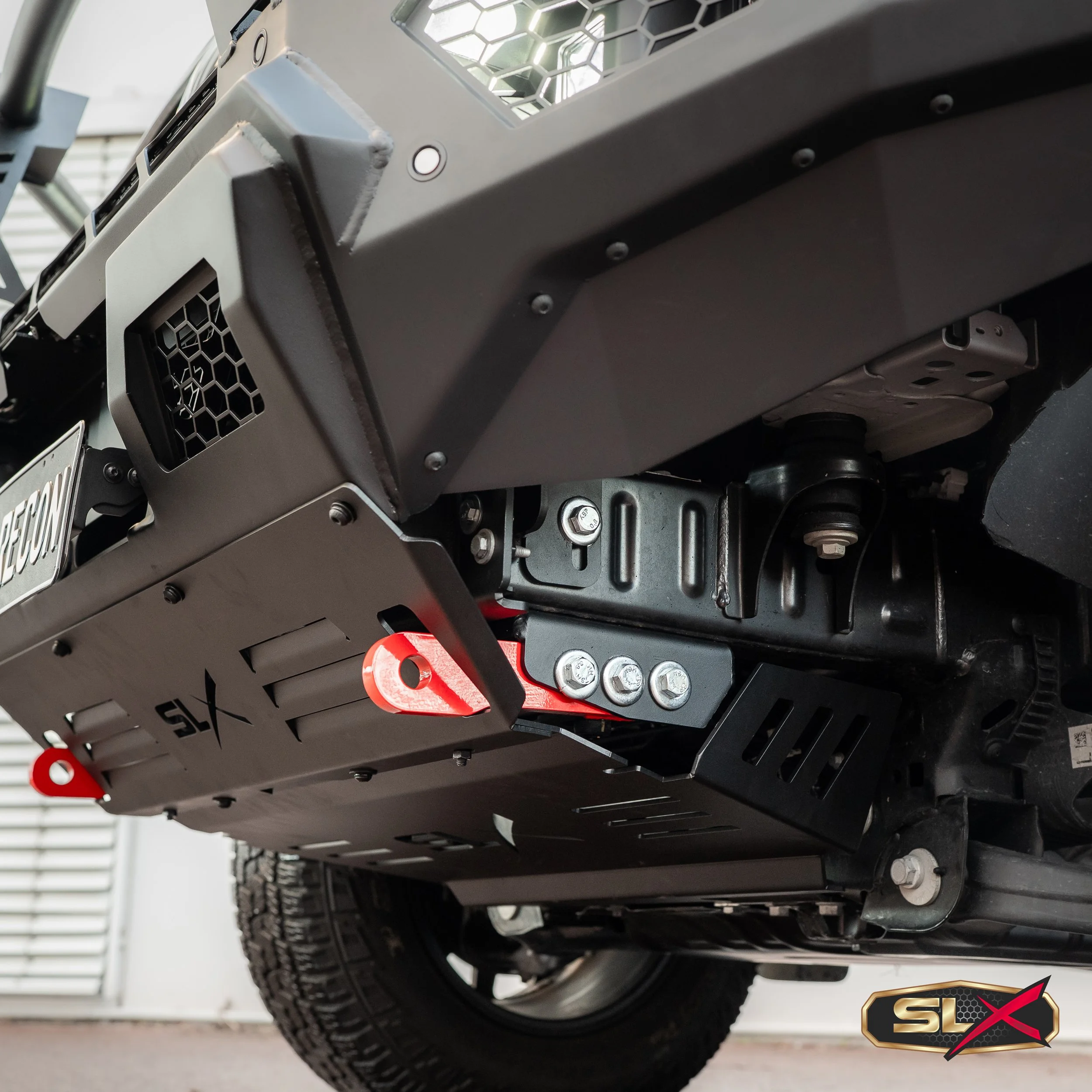Close-up of the underside of a vehicle, showing a black skid plate, metal bolts, brackets, and parts of a tire, with a logo in the bottom right corner that reads SLYX.