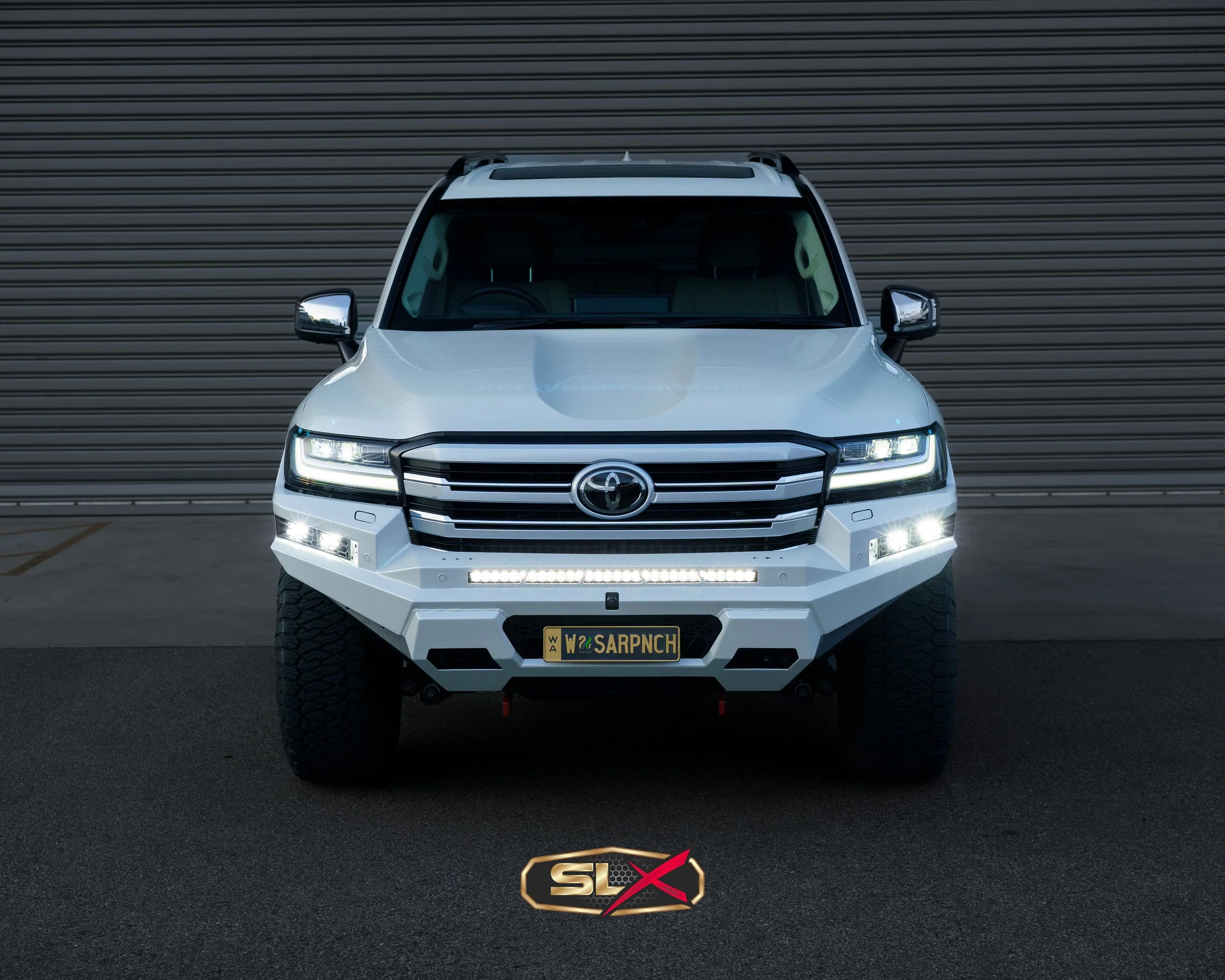 White off-road SUV with black grille and bumper, LED headlights, and large tires, parked facing forward in front of a metal garage door.