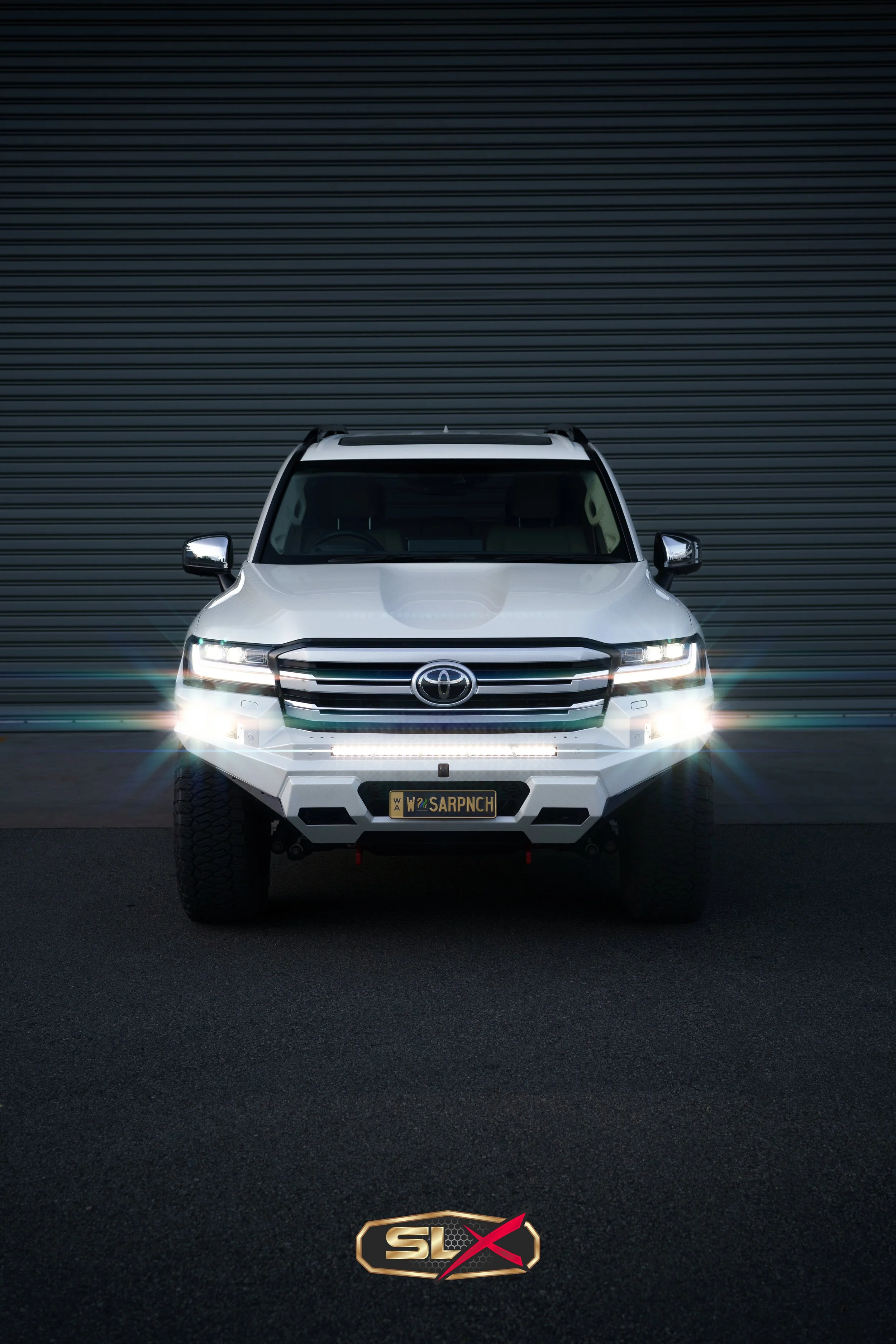 Front view of a white Toyota SUV with headlights and daytime running lights on, parked in front of a dark, ribbed metal door, with a gold decal reading 'SLX' on the pavement.