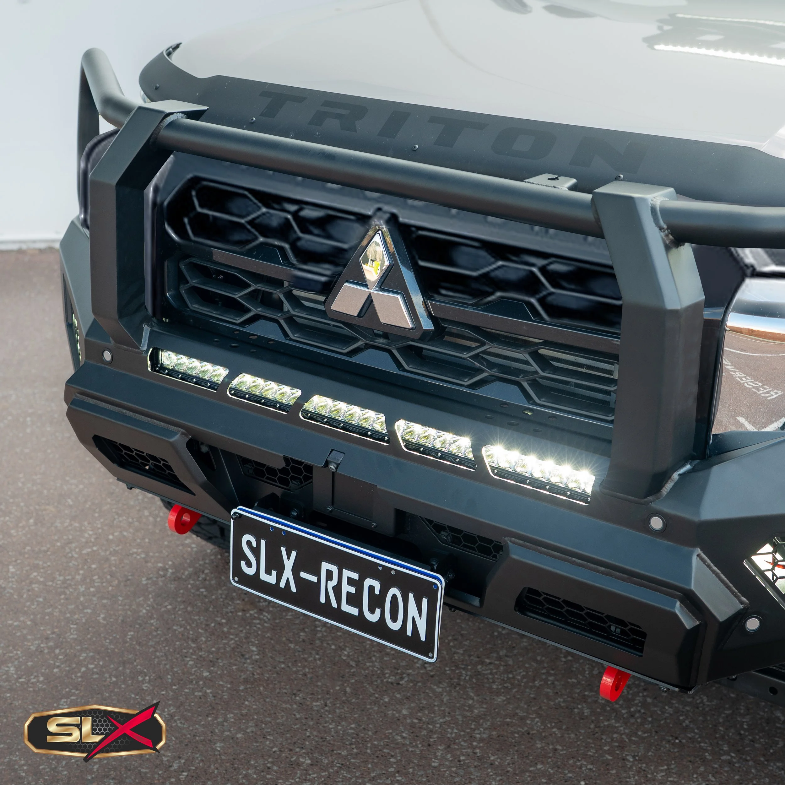 Close-up of the front of a rugged off-road vehicle with a black grille, Mitsubishi emblem, LED lights below the grille, a license plate reading SLX-RECON, and a black bumper with red tow hooks.