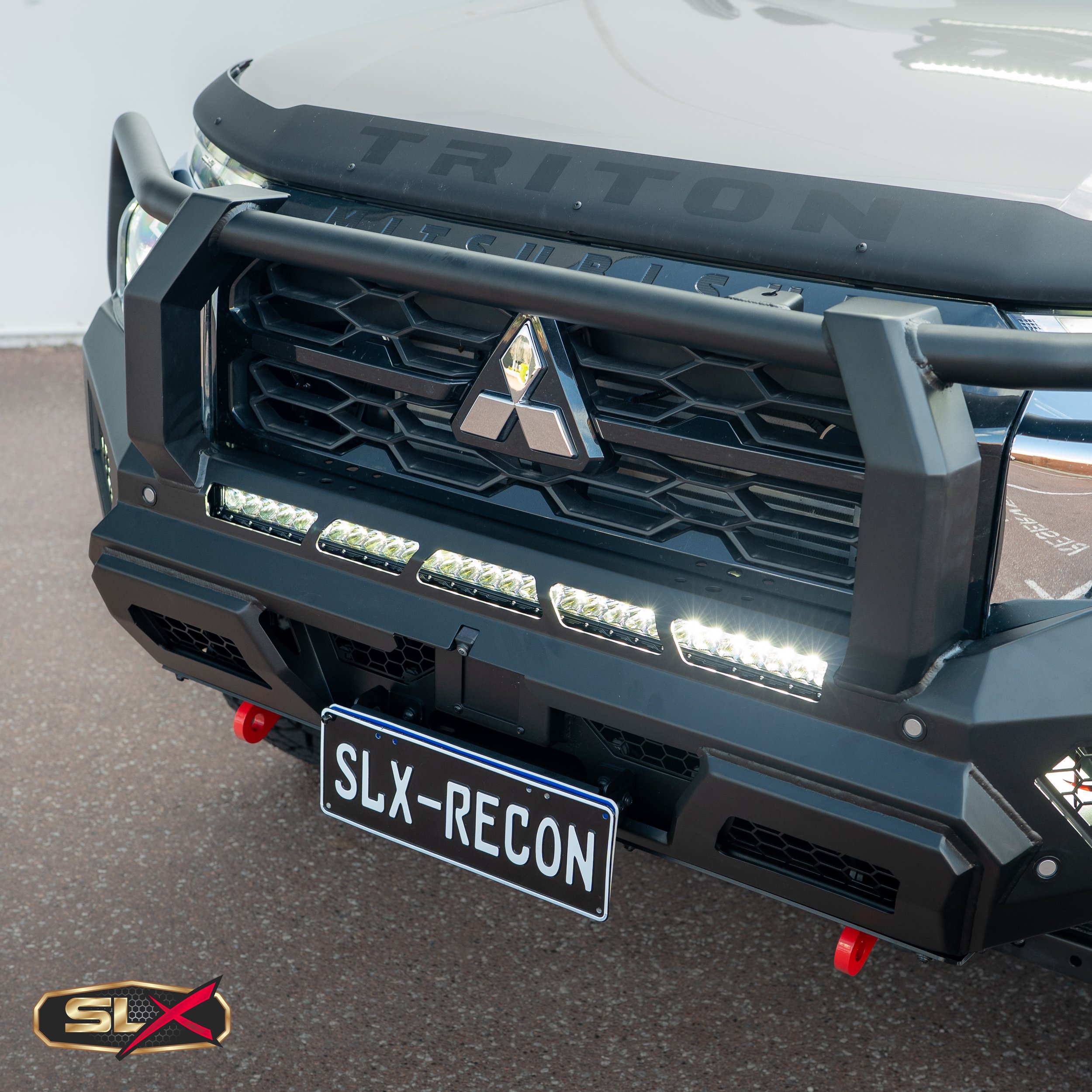Close-up of rugged black off-road vehicle front with Mitsubishi logo, LED light bar, and custom SLX-RECON license plate.