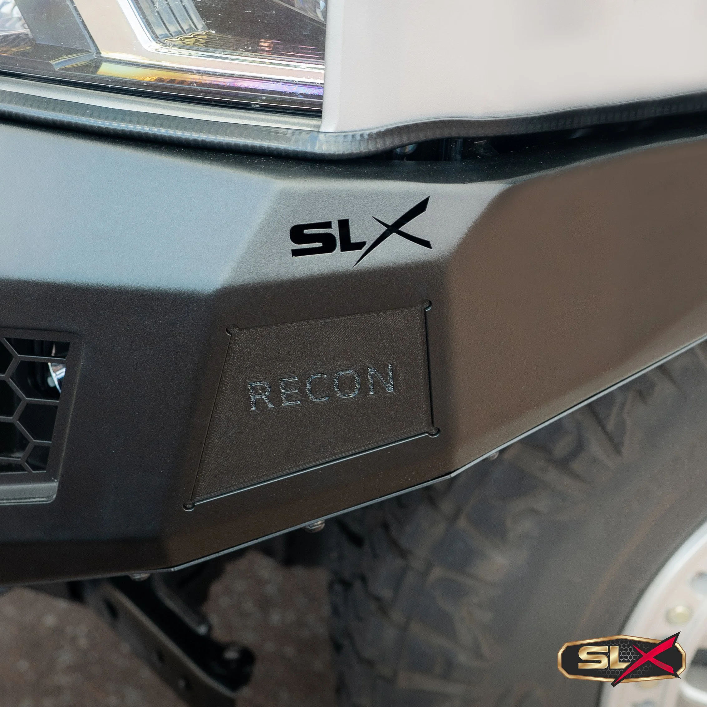 Close-up of a vehicle's front end showing a headlight, a black badge with the letters "SLX" and a stylized X, and a small black panel with the word "RECON" on it.