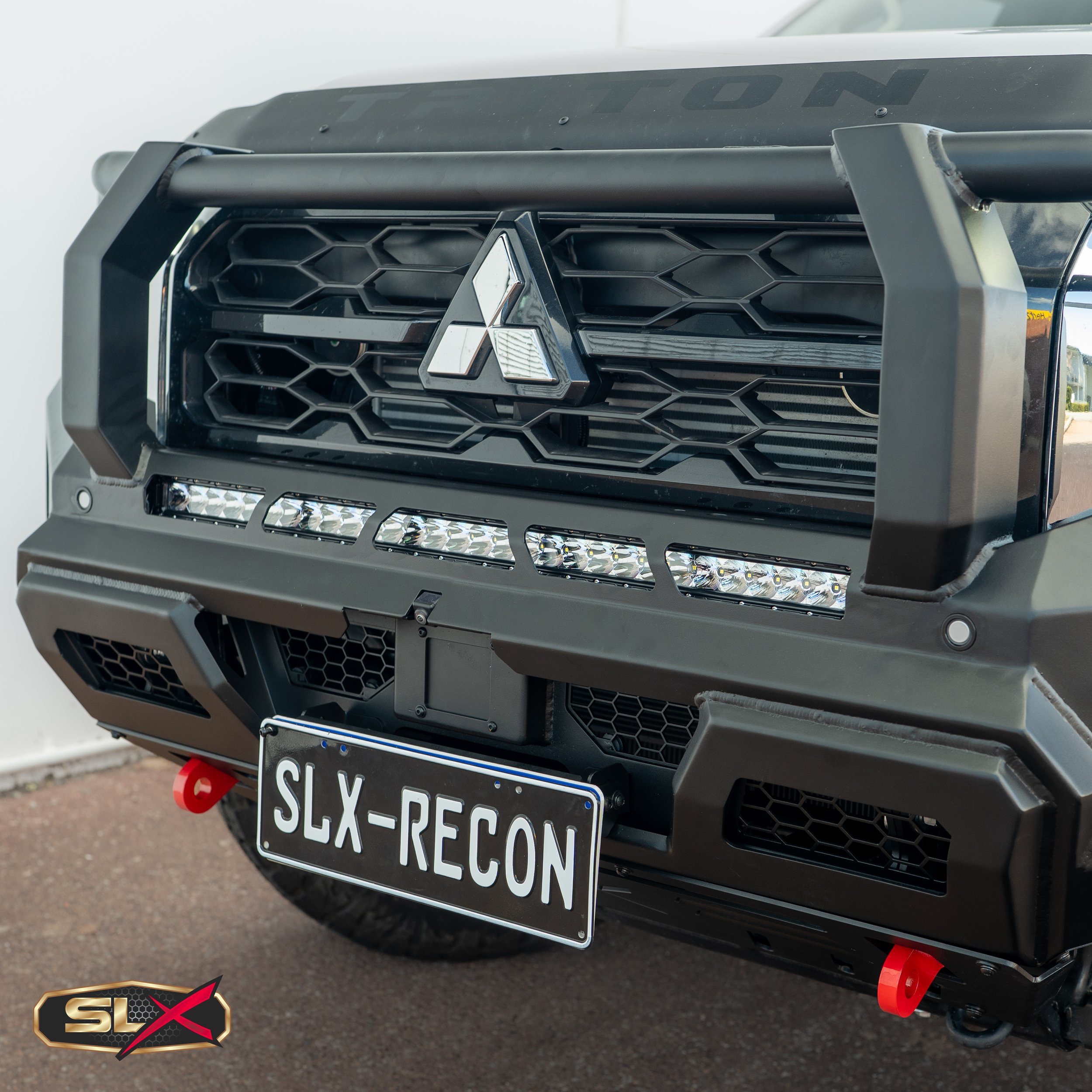 Close-up of a black off-road vehicle's front grille with a Mitsubishi logo, LED light bar, and custom black bumper with red hooks, license plate reading SLX-RECON, and SLX logo in the bottom left corner.