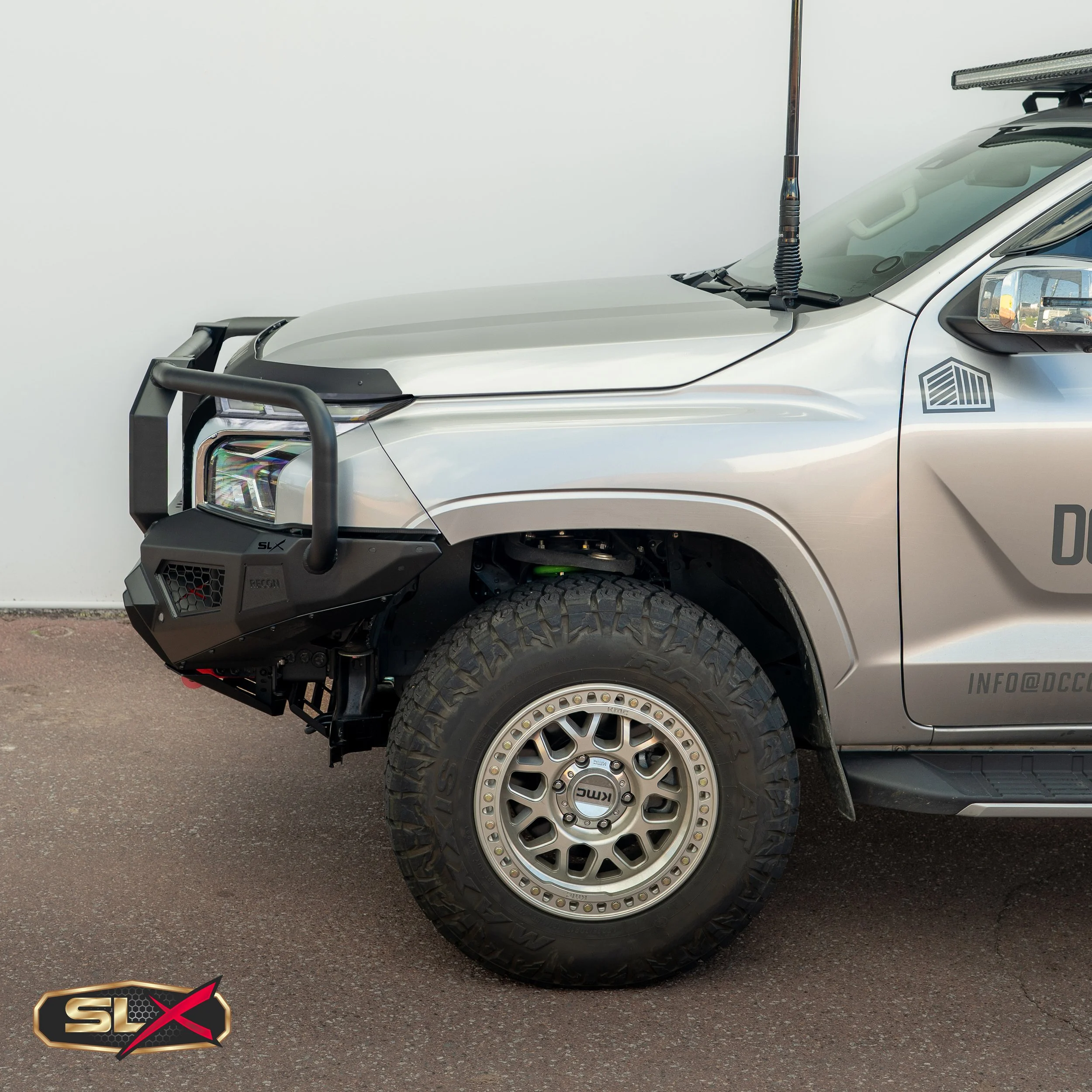 Close-up of the front part of a silver off-road vehicle with a rugged tire, black front bumper with protective guard, and a mounted antenna on the hood.