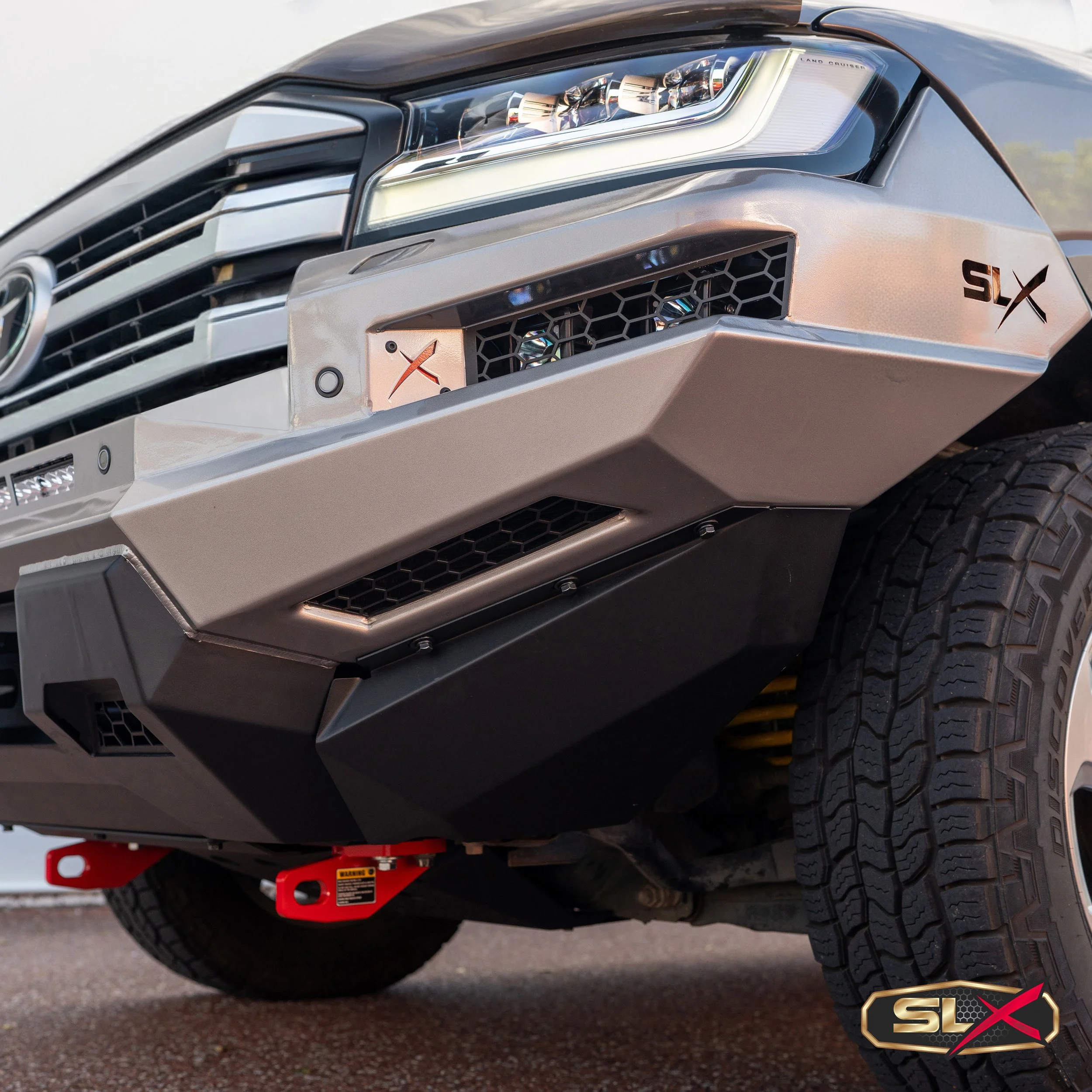 Close-up of the front of a modern pickup truck, showing the headlight, grille, and bumper with SLX logos. The truck has large tires and a red tow hook below the bumper.