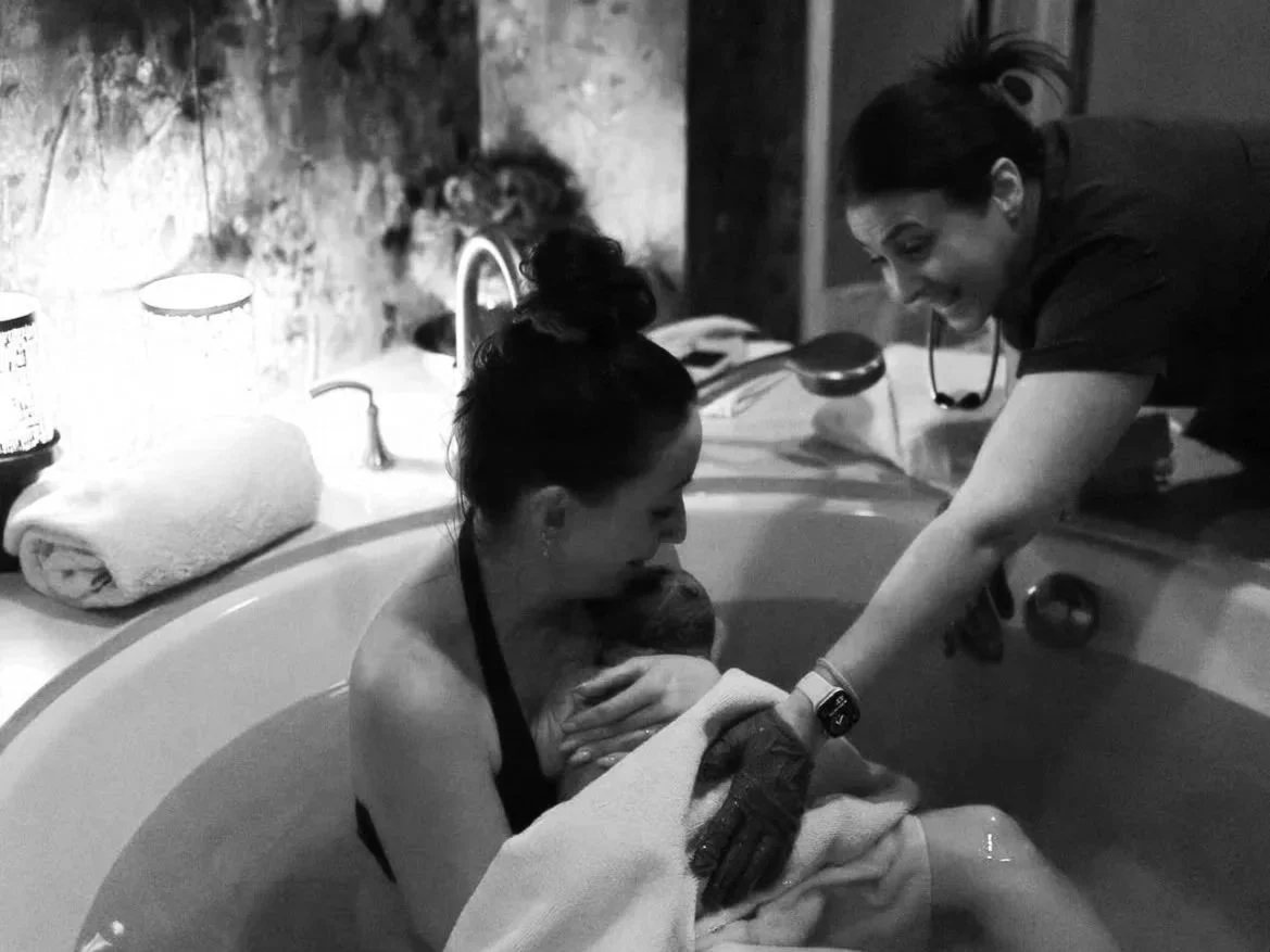 A woman holding a newborn baby in a bathtub while another woman assists and smiles.