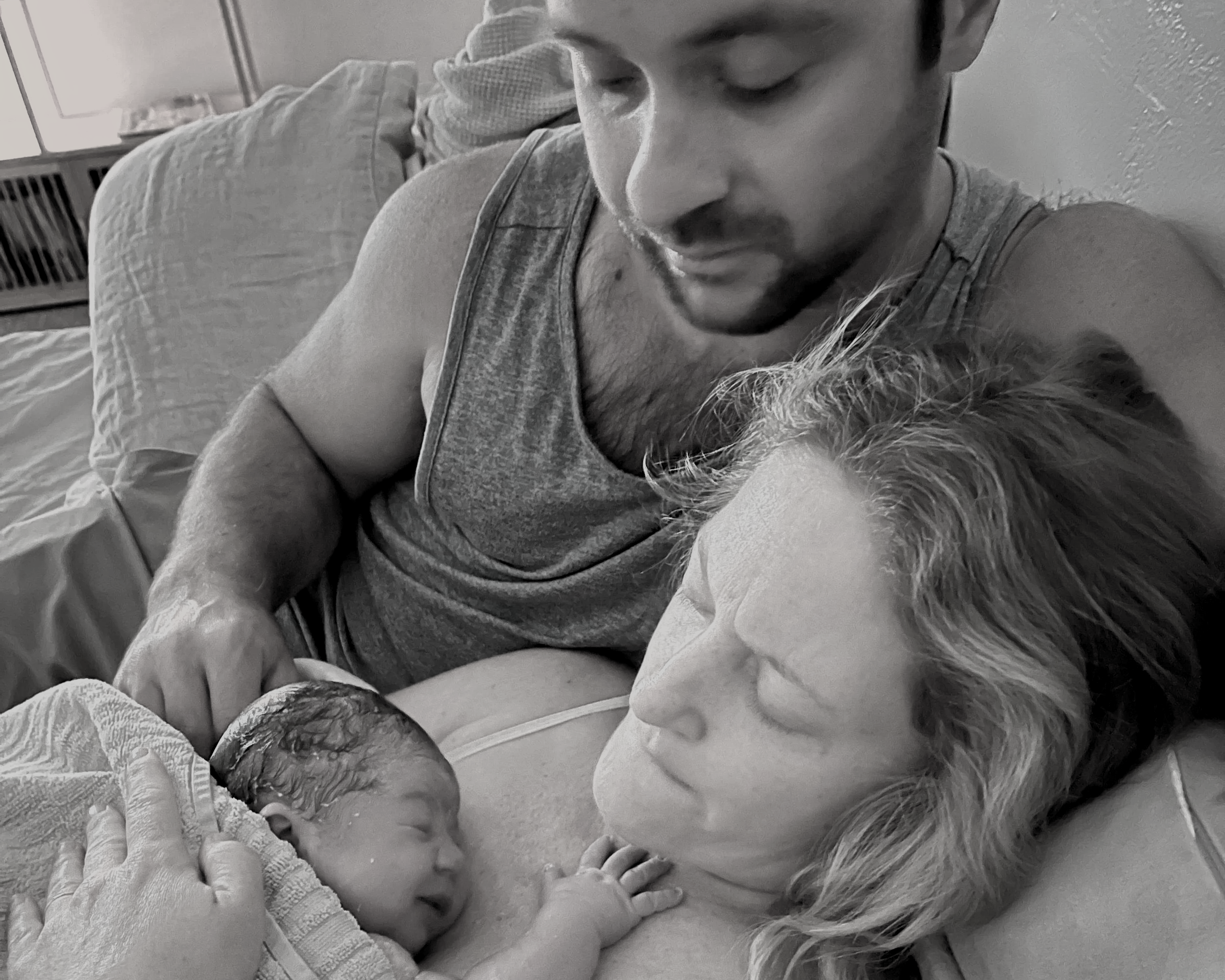 A man, woman, and newborn baby atnhome with the mother lying in bed and the man standing close by, looking at the baby.