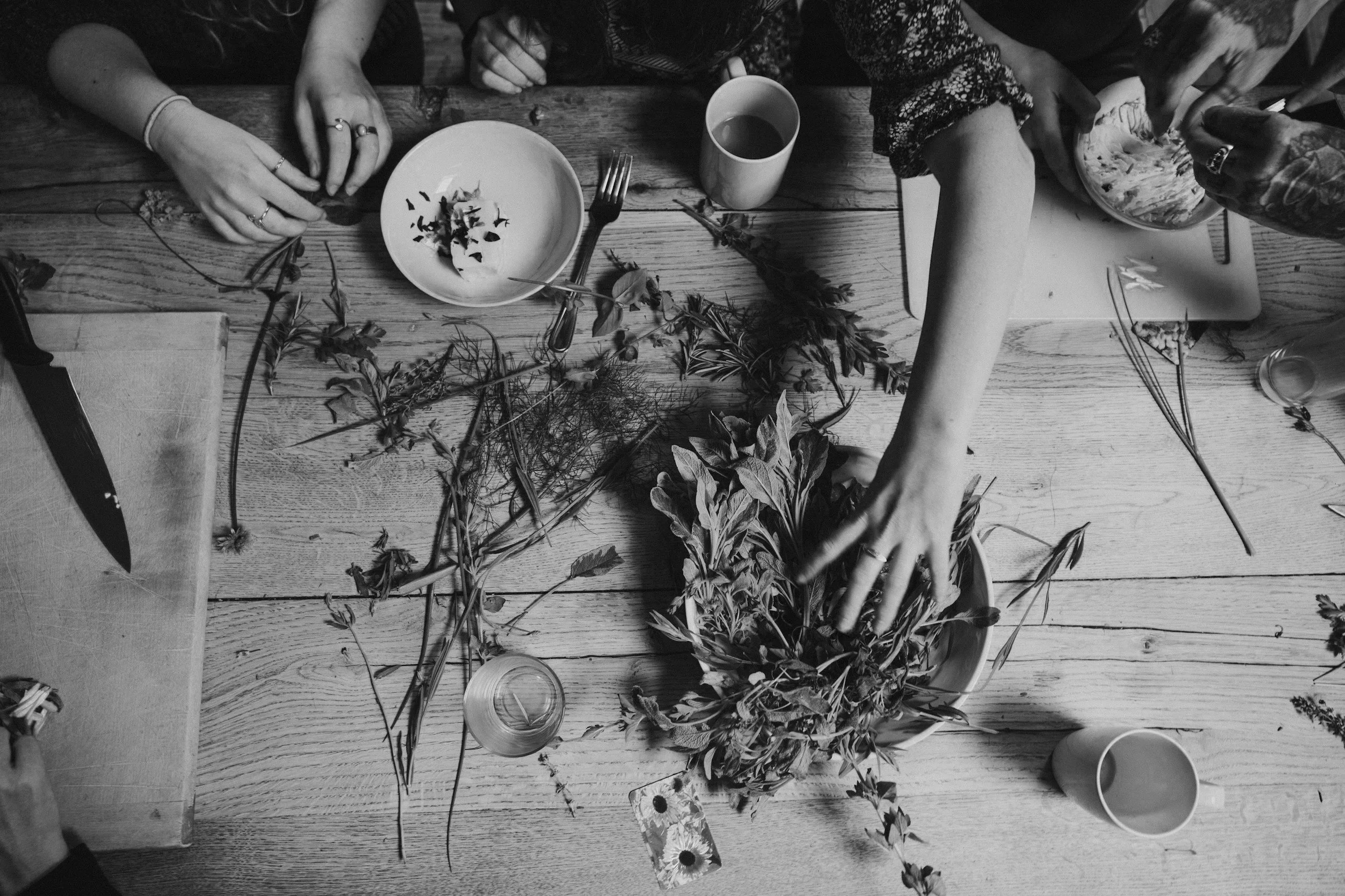 People gathered around a wooden table, arranging flowers and herbs, with dishes, cups, utensils, and a knife visible on the table.