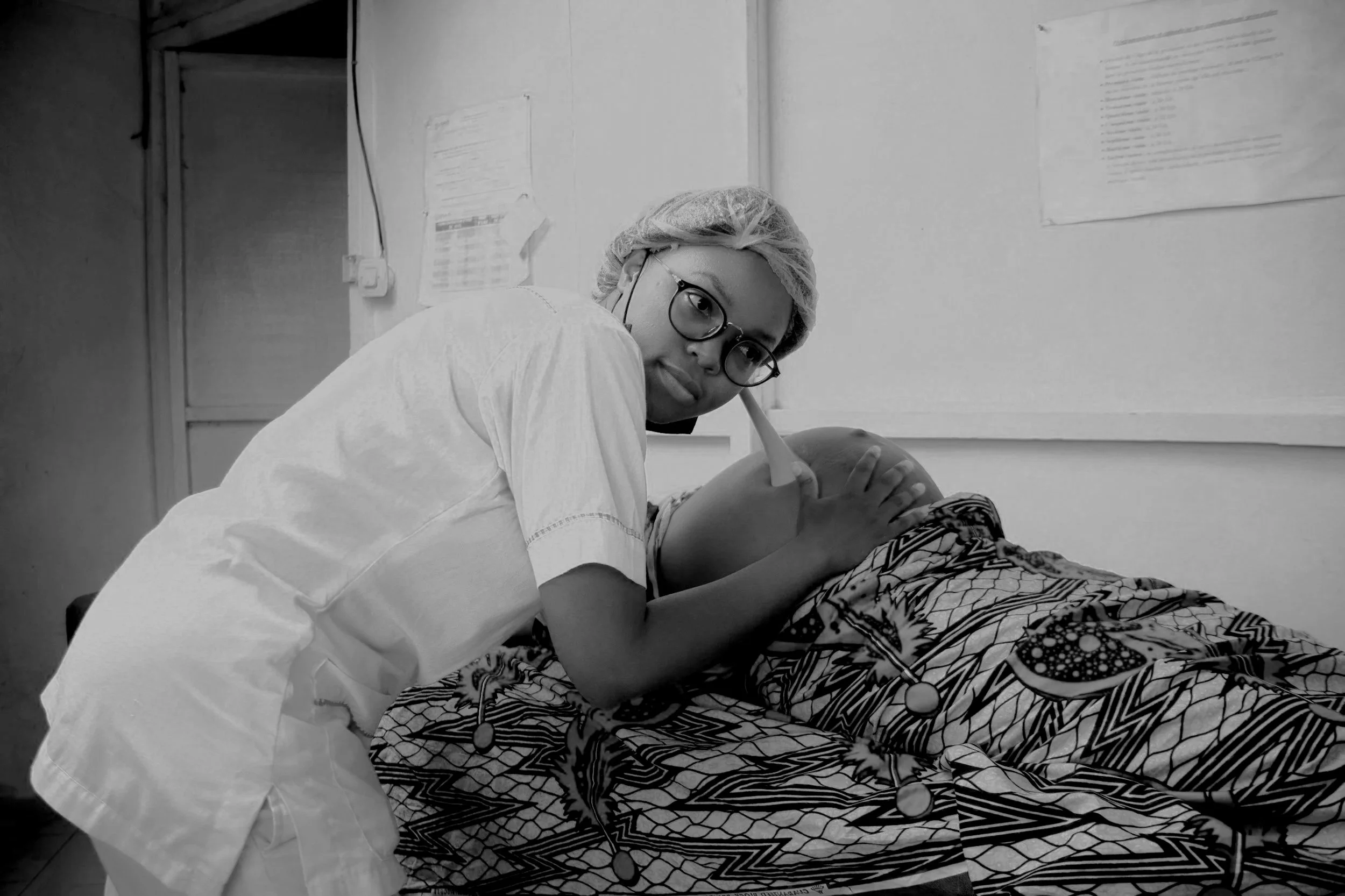 A nurse in scrubs, hairnet, and glasses cares for a newborn in a hospital room, with medical charts on the wall.