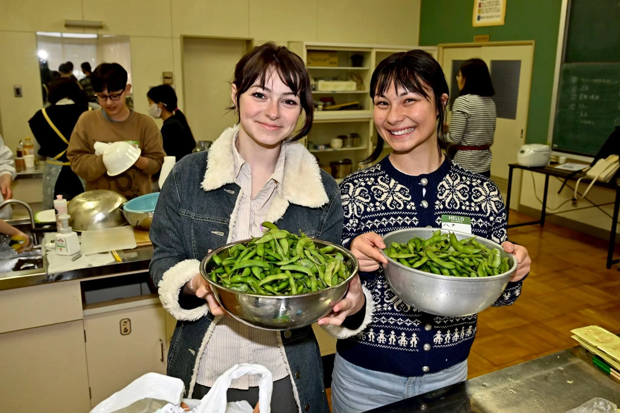Two women smiling and holding large bowls of edamame at a community event, with other people in the background preparing food in a kitchen setting.