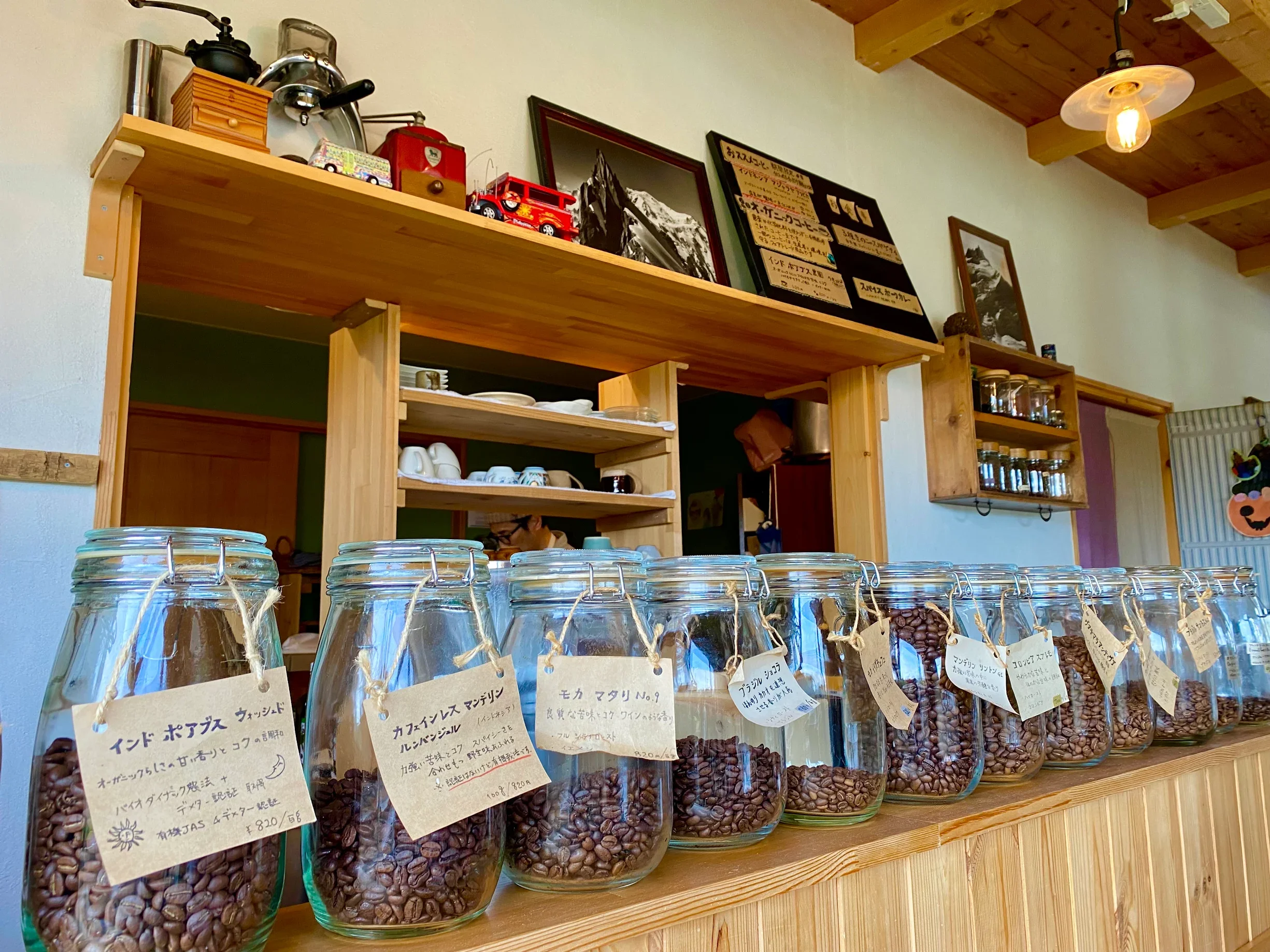 A row of glass jars filled with roasted coffee beans on a wooden shelf. Each jar has a handwritten label with descriptions in Japanese. In the background, there are shelves with dishes, framed photographs, and other decorative items on the wall, with a warm wooden ceiling and hanging light bulb.