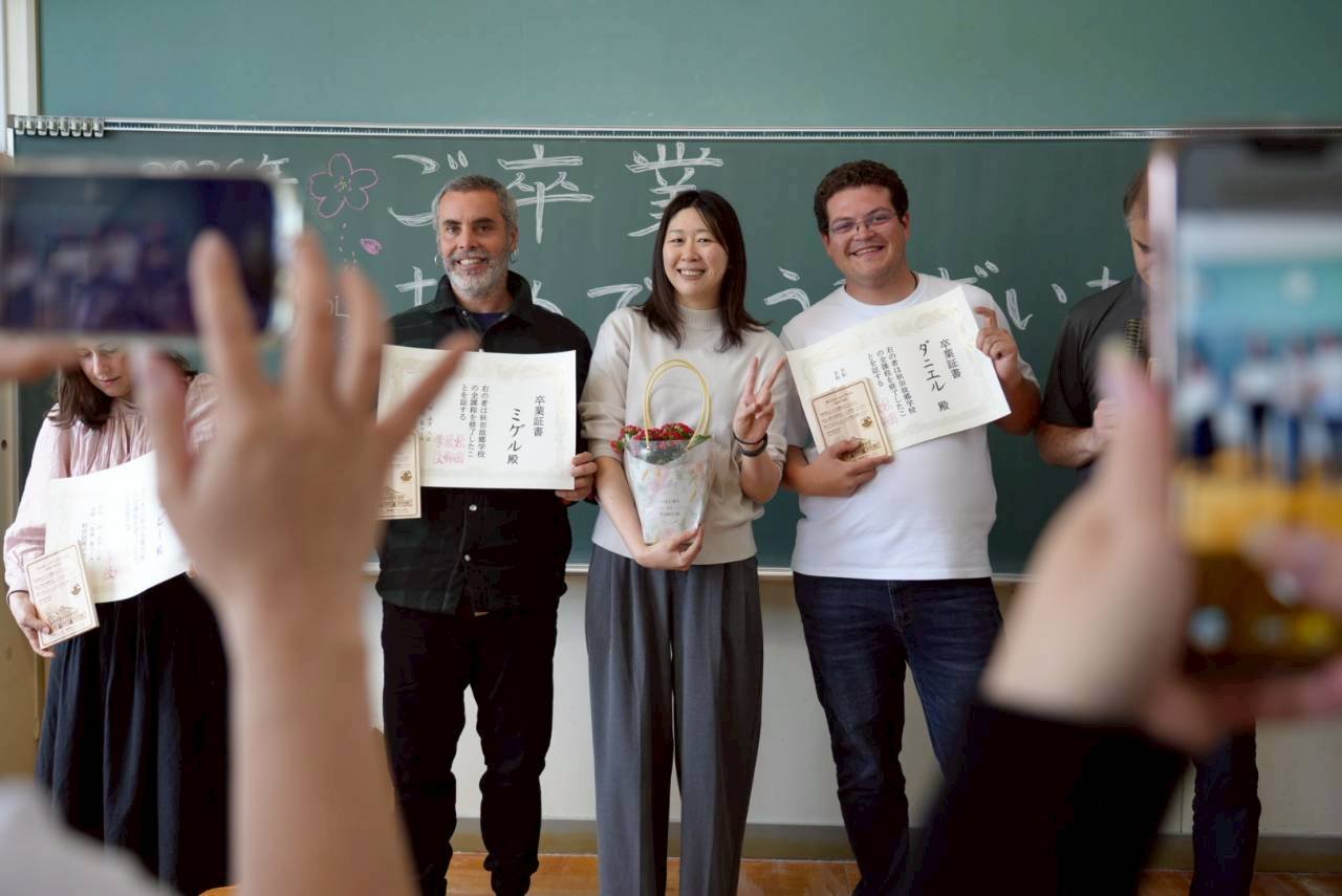 People taking a photo of three individuals in front of a blackboard, with certificates and a gift basket, celebrating an achievement or event in a classroom.