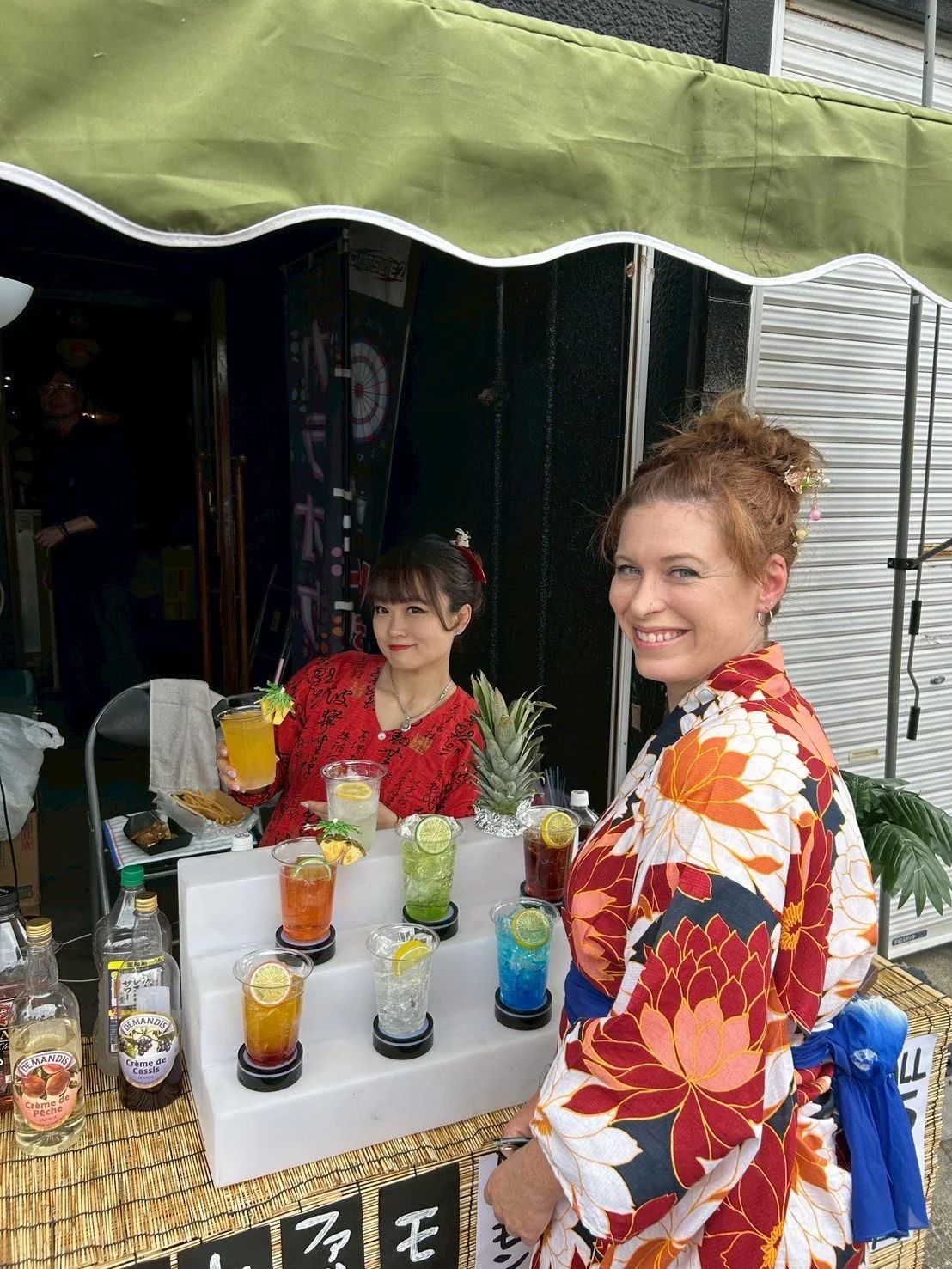 Two women at a drink stand, with colorful cocktails garnished with lime slices and small umbrellas, and a pineapple on display. The woman on the right is smiling and wearing a floral-patterned kimono, while the woman on the left, with dark hair and a red outfit, is holding a yellow cocktail and winking.